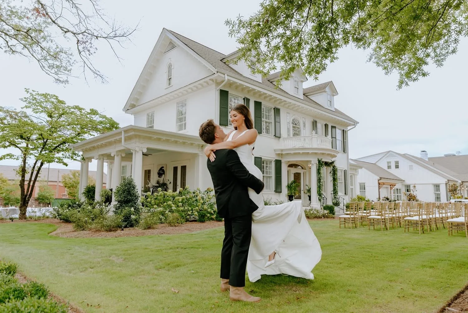 A couple in wedding attire standing on a lawn, with the man lifting the woman in front of a large white house with green shutters.