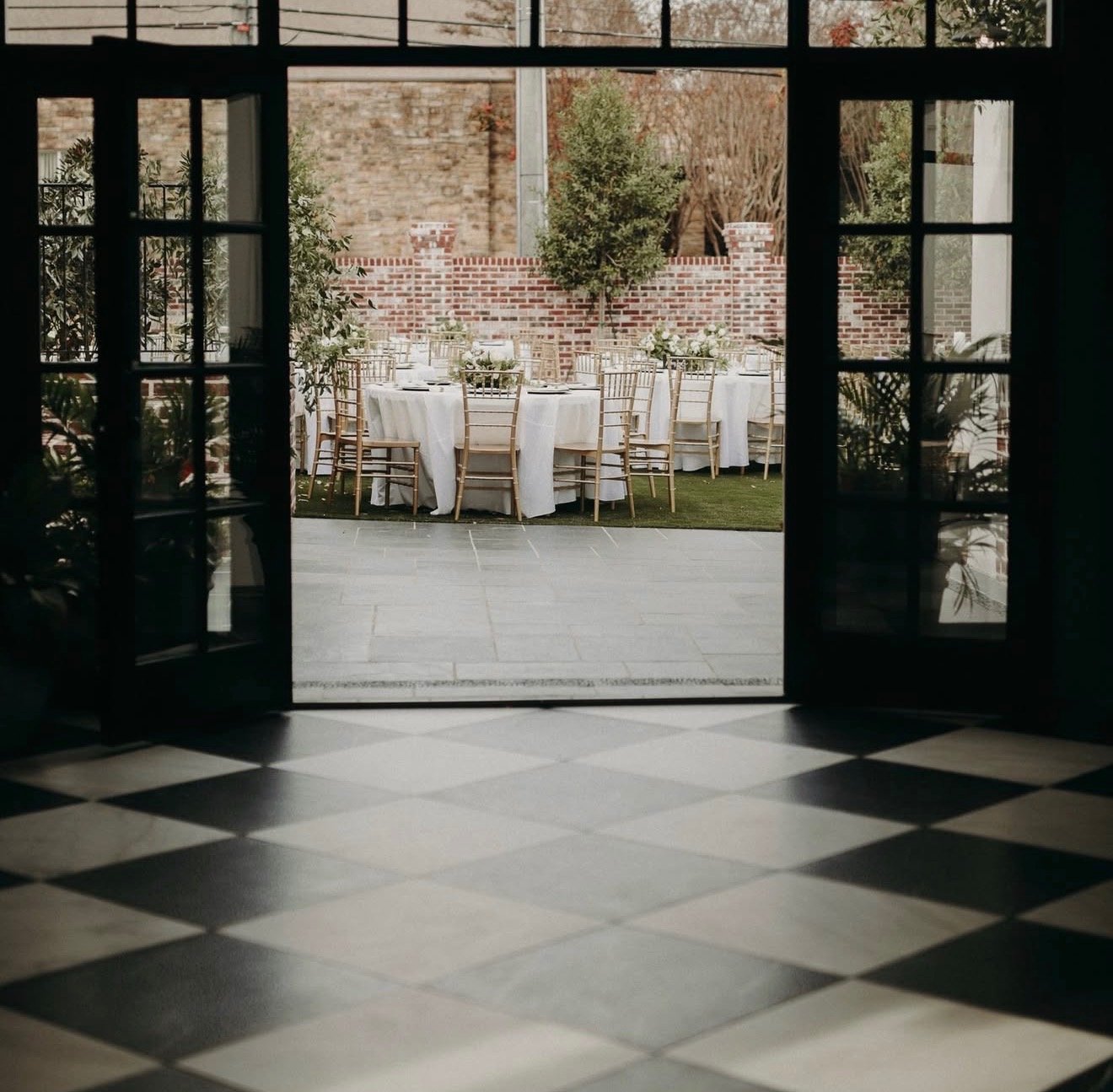 View through glass doors showing outdoor event setup with round tables, white tablecloths, and gold chairs, surrounded by brick wall and greenery.
