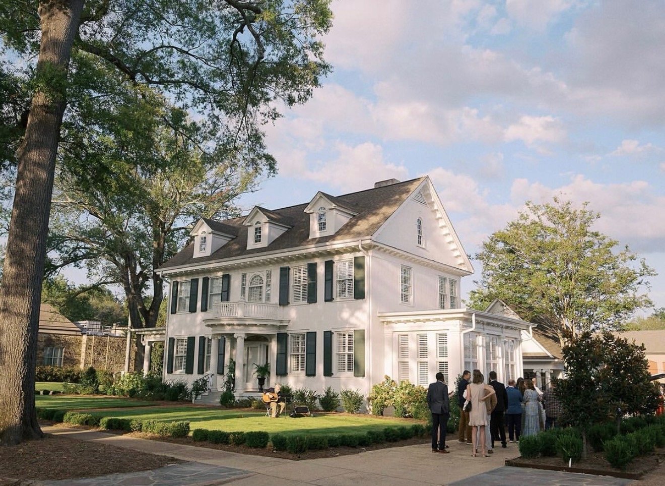 A group of people gathered outside a large white Victorian-style house with dark shutters on a sunny day.