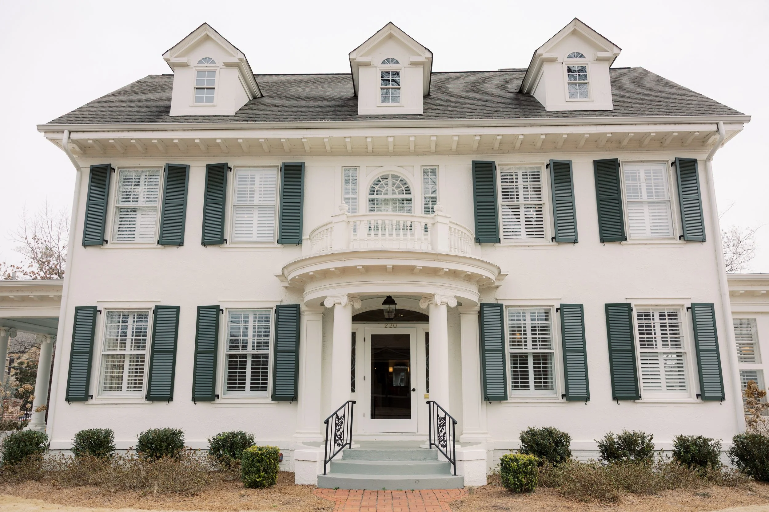 White three-story house with black shutters, dormer windows, a small balcony above the front door, and a black lantern light hanging at the entrance.