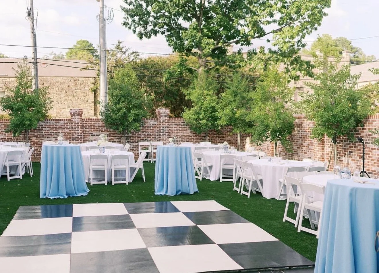 Outdoor event setup with round tables covered in white and blue tablecloths, surrounded by white folding chairs, on artificial grass with a brick wall and trees in the background and a black-and-white checkered dance floor in the foreground.