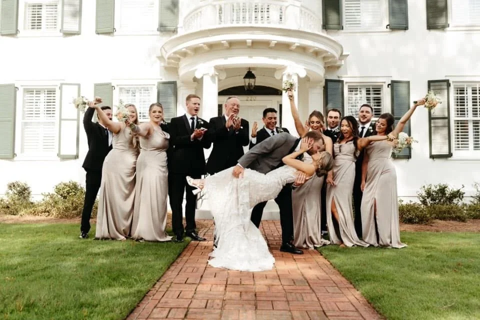 Wedding party outside a large white house with blue shutters. Bride and groom are kissing, with the groom dipping the bride. Six women in matching gray dresses and four men in black suits surround them, celebrating and holding bouquets.