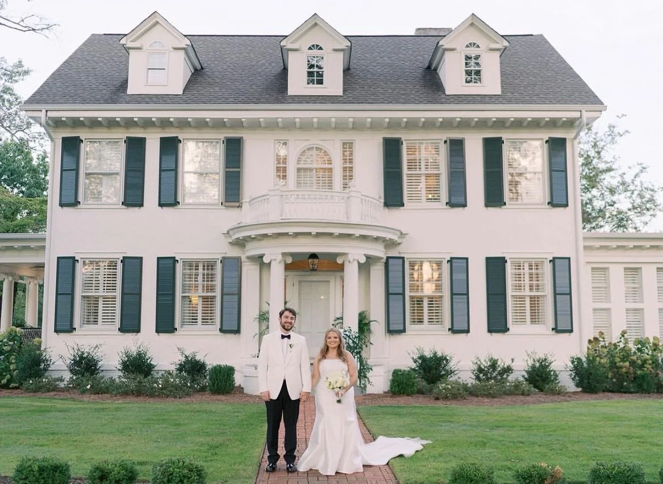 A newlywed couple standing in front of a large white house with black shutters on a well-manicured lawn.
