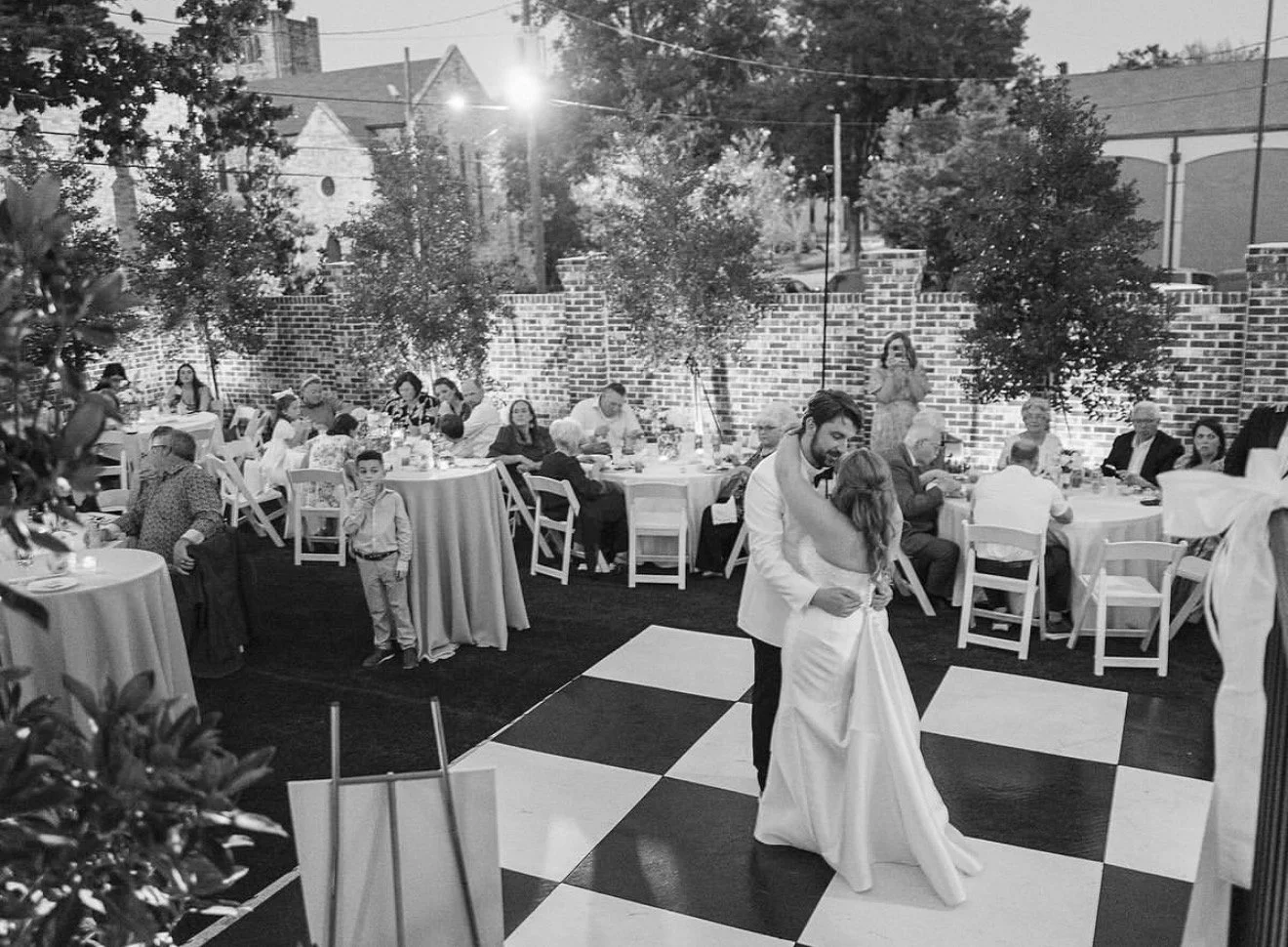 Couple dancing at a wedding reception on a checkered dance floor, with guests seated at round tables with white tablecloths in the background, outdoor setting with brick wall, trees, and string lights.