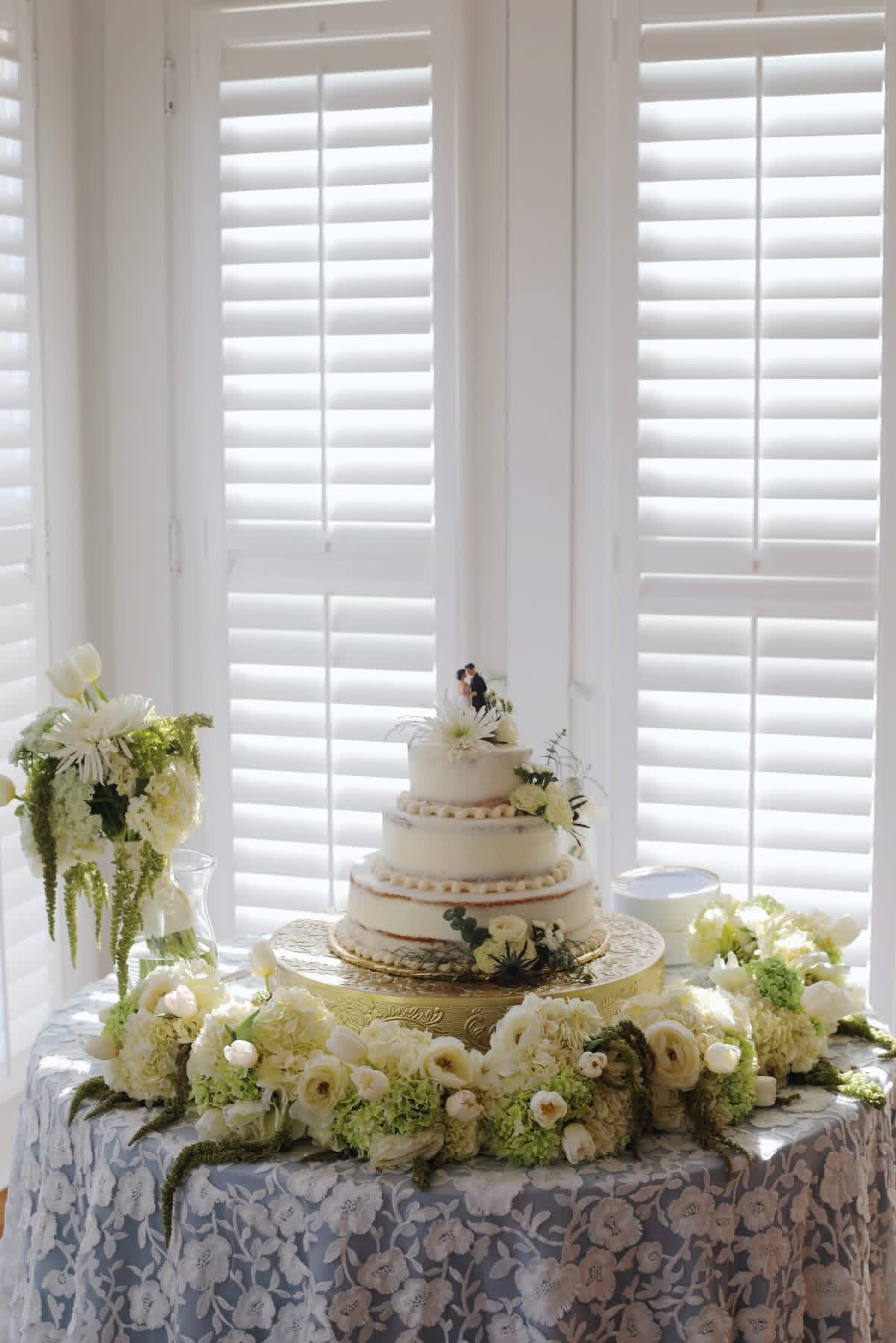 A three-tier wedding cake decorated with white flowers and greenery, topped with a bride and groom figurine, placed on a round, gold-colored stand surrounded by white flowers on a table with a lace tablecloth. Behind the table are white shutters.