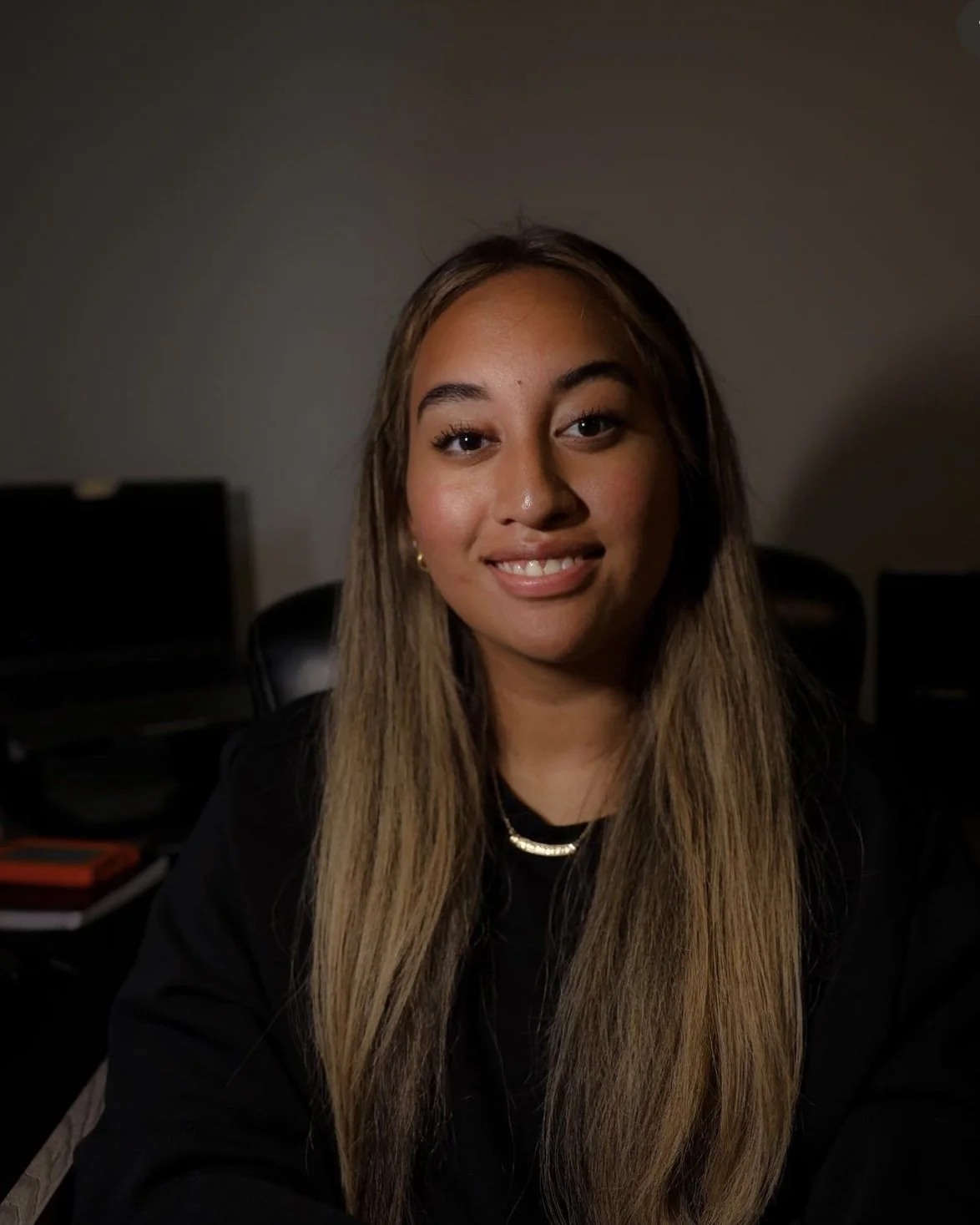 A young woman with long, light brown hair smiling at the camera in a dimly lit room.