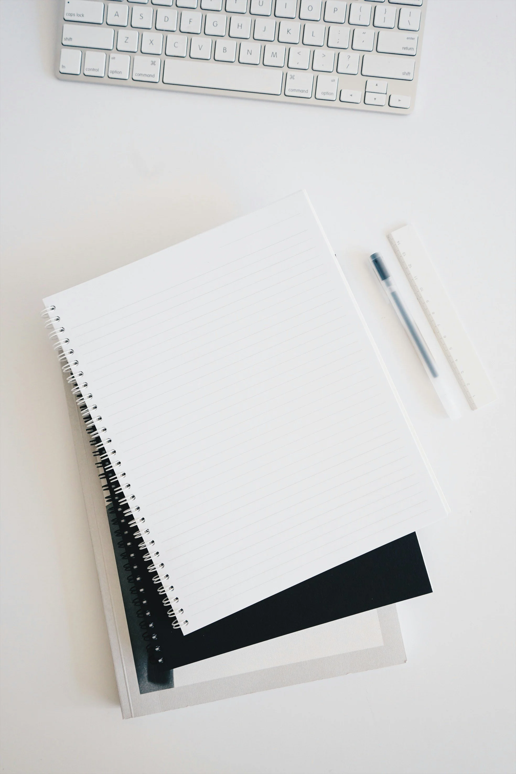 Stationary items on a white desk, including a spiral notebook, a black notebook underneath, a grey notebook underneath, a pen, a ruler, and a computer keyboard.