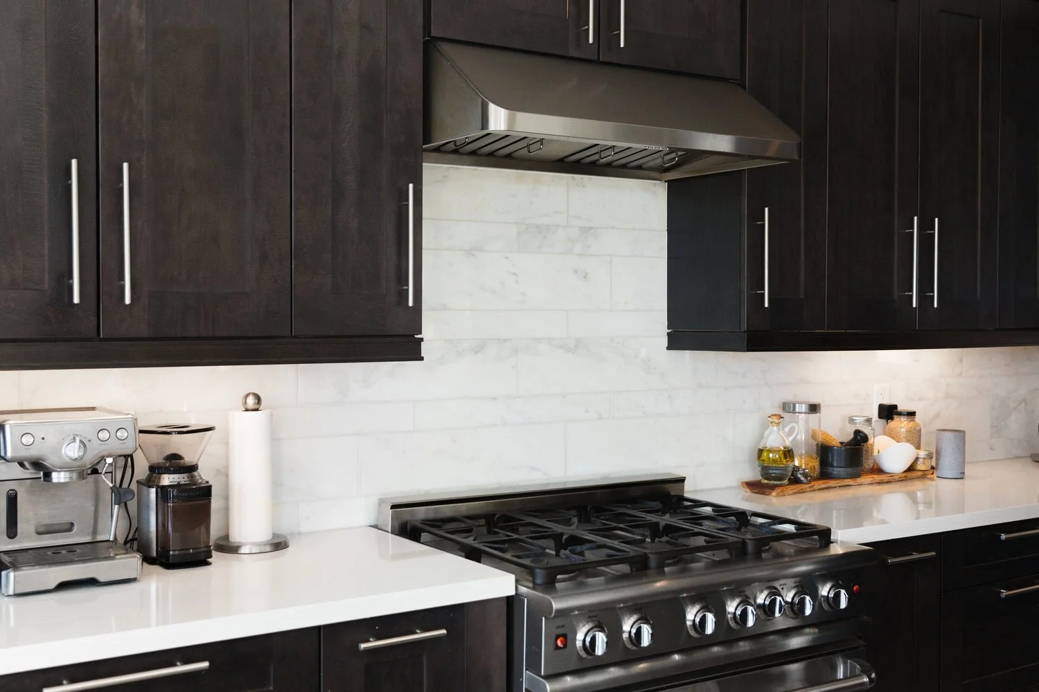 Modern kitchen with dark wood cabinets, stainless steel range hood, and a white countertop. Kitchen appliances include a coffee maker, and a grinder, with various kitchen jars and oils on a wooden tray.