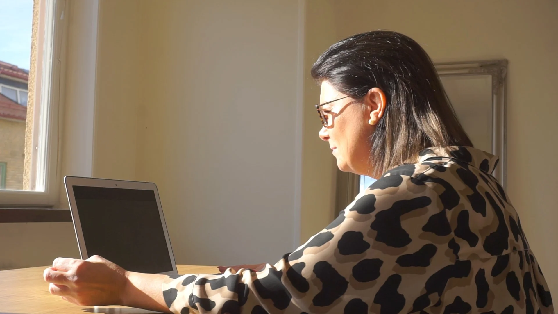 A woman with dark hair and glasses sitting at a table, looking at an electronic device, during daytime with sunlight coming through a window.