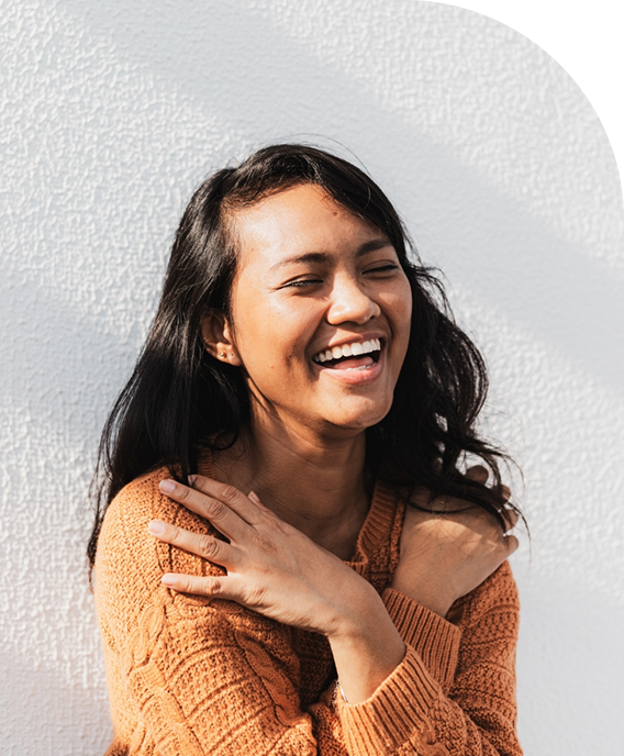 A woman with long dark hair smiling and hugging herself against a light textured wall.