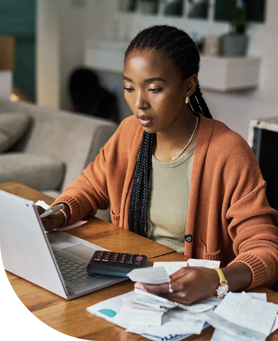 An African-American woman reviewing financial planning documents at a wooden table with a laptop, calculator, receipts, and papers in a modern living room.