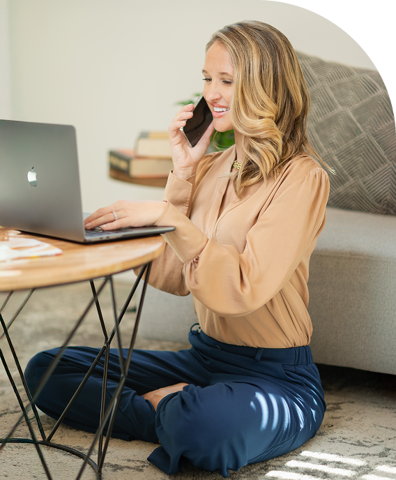 Theresa Peterson sitting cross-legged on the floor talking with a client while working on her laptop, representing approachable financial guidance and personalized planning.