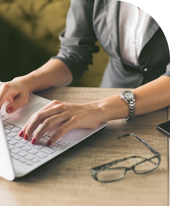 Close-up of a woman's hands typing on a laptop keyboard, with glasses and a smartphone on a wooden desk.