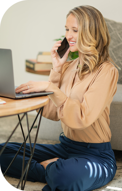 Theresa Peterson sitting cross-legged on the floor talking with a client while working on her laptop, representing approachable financial guidance and personalized planning.