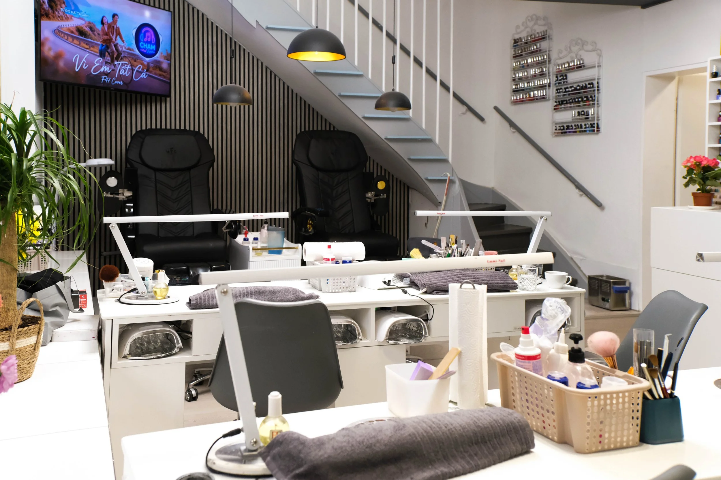 Ein Nagelstudio mit Behandlungsstühlen, einem Verkaufsregal für Nagellacke, einem Fernseher an der Wand, mehreren Arbeitsstationen mit Werkzeugen und Pflegeprodukten auf Tischen, sowie Pflanzen und Dekorationsgegenständen