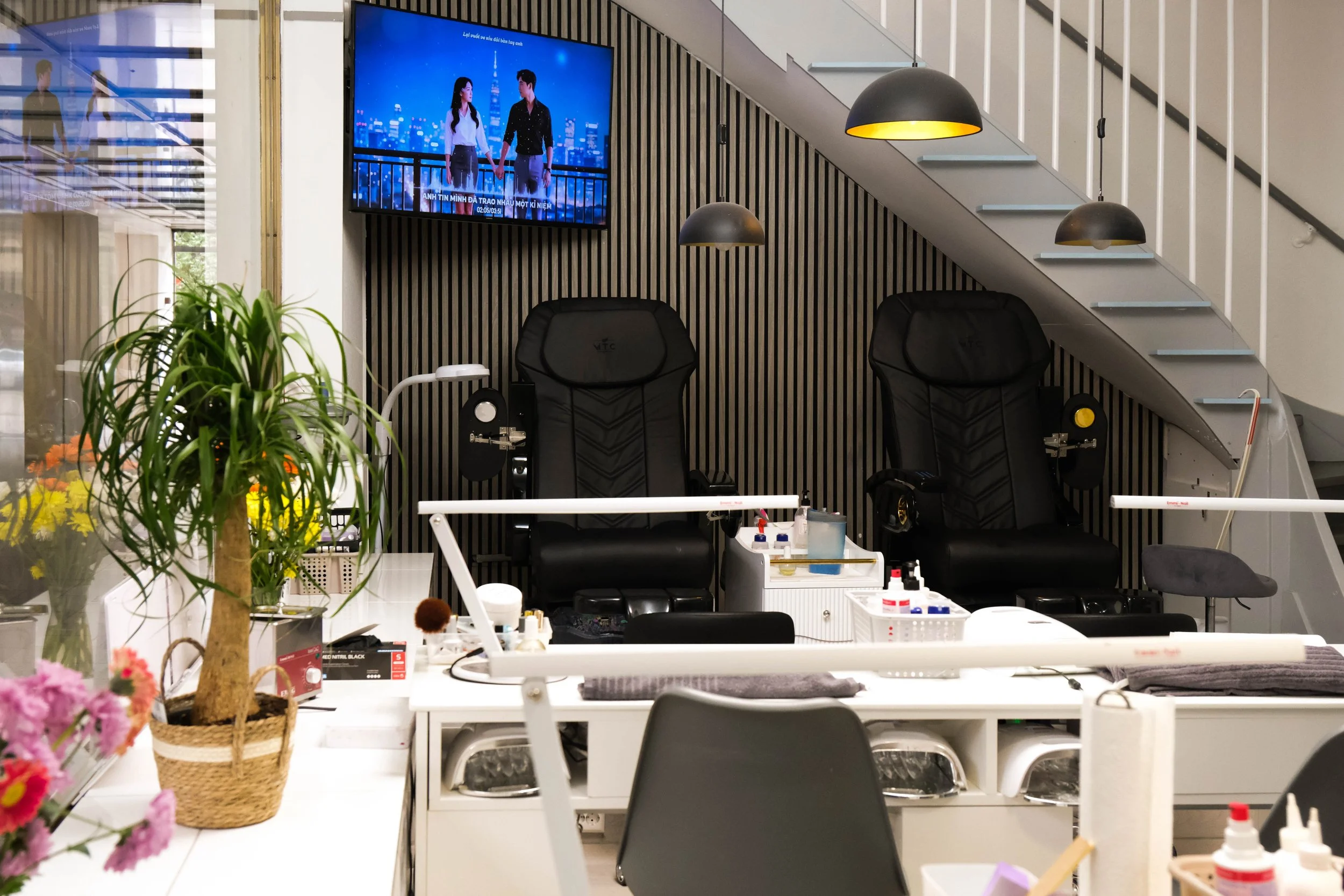 Ein modern eingerichteter Raum mit Laserdruckstationen, Pflanzen und Schreibtischen, Wasserpistolen, Nagellack und Make-up-Produkten auf den Tischen, und einem Fernseher an der Wand, der zwei Personen auf einem Balkon zeigt.