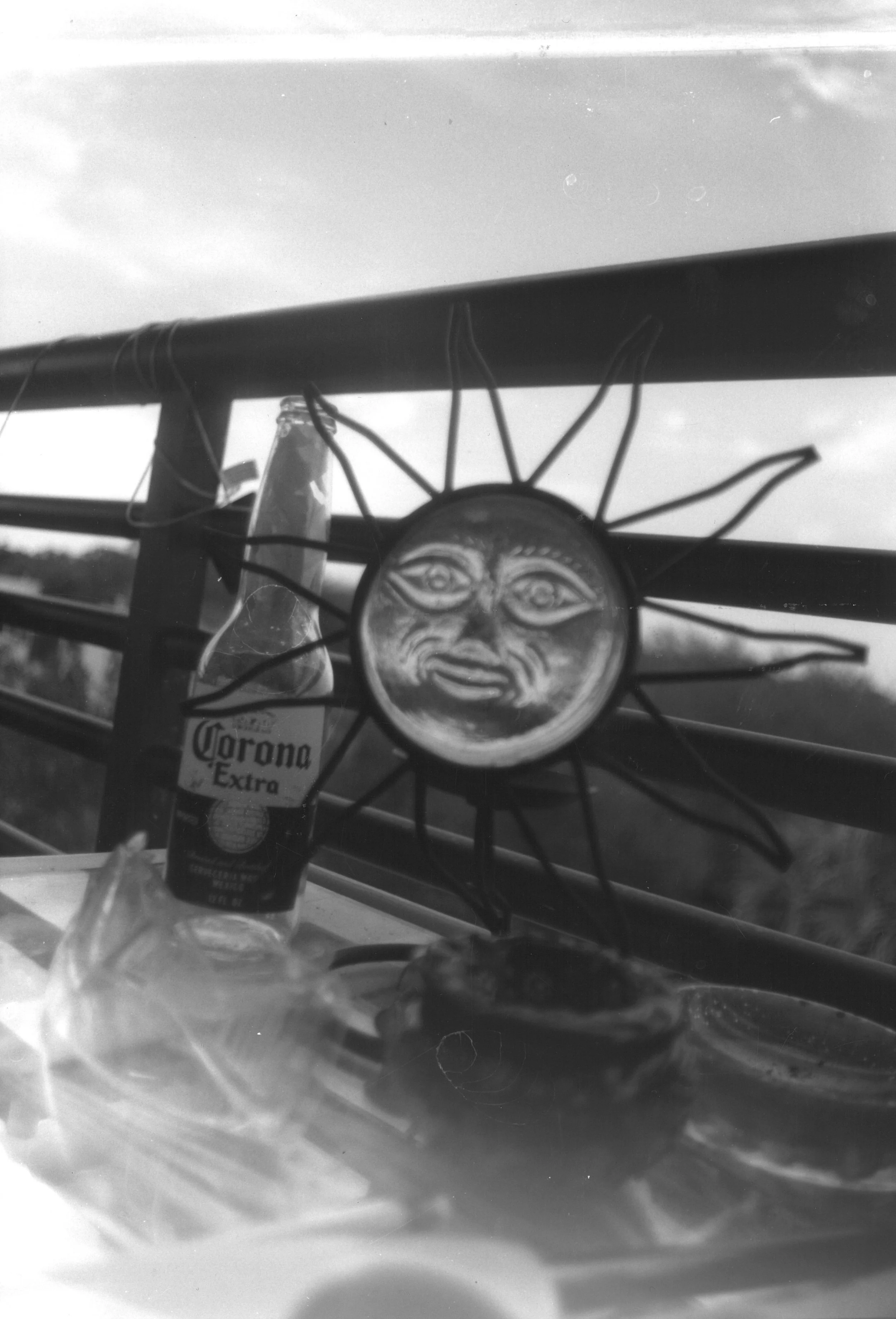 A decorative sun-shaped metal wall hanging with a face, hanging on a railing outdoors, next to a bottle of Corona Extra beer.