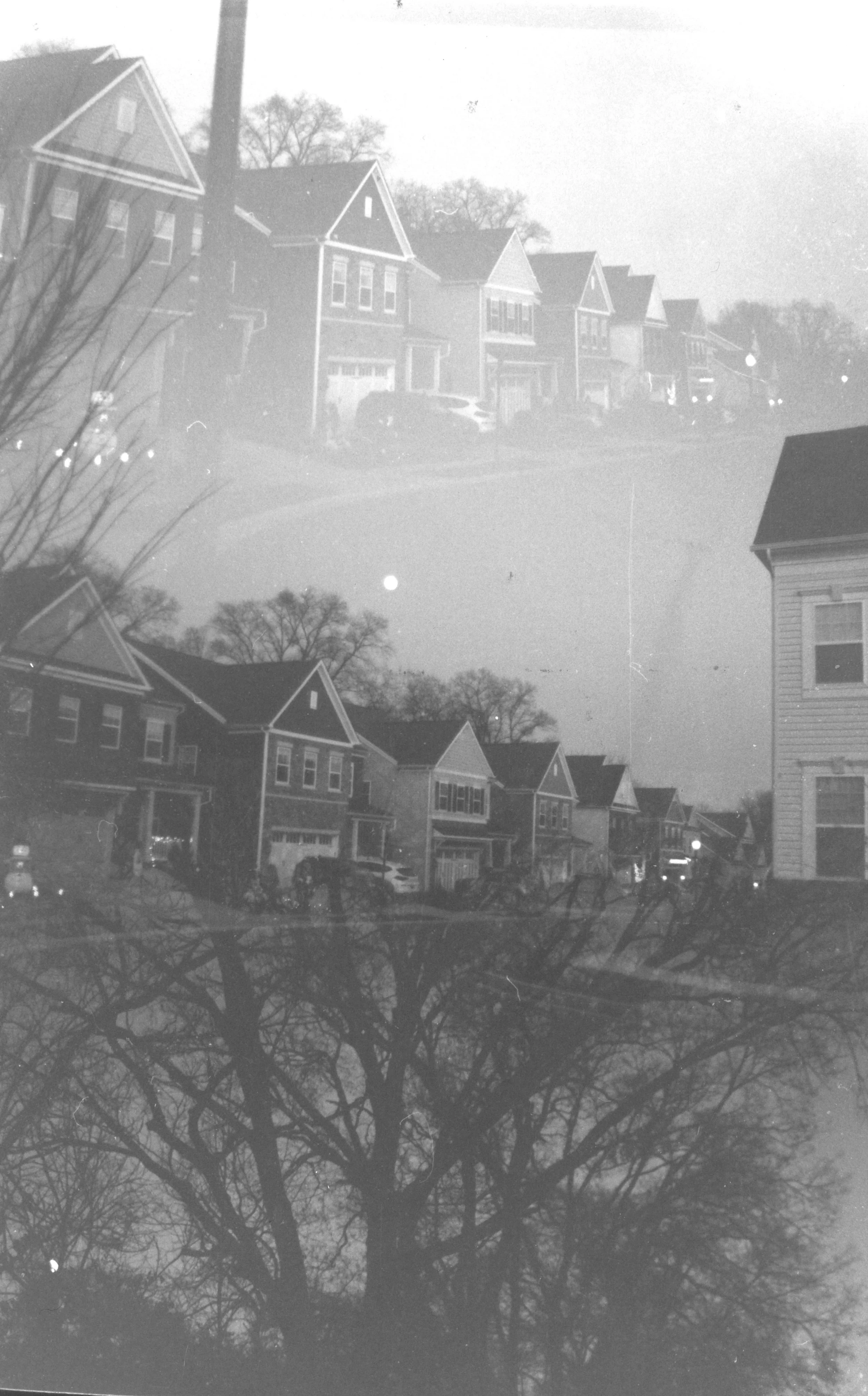 A double exposure black and white photo of a residential street with houses, trees, parked cars, and streetlights.