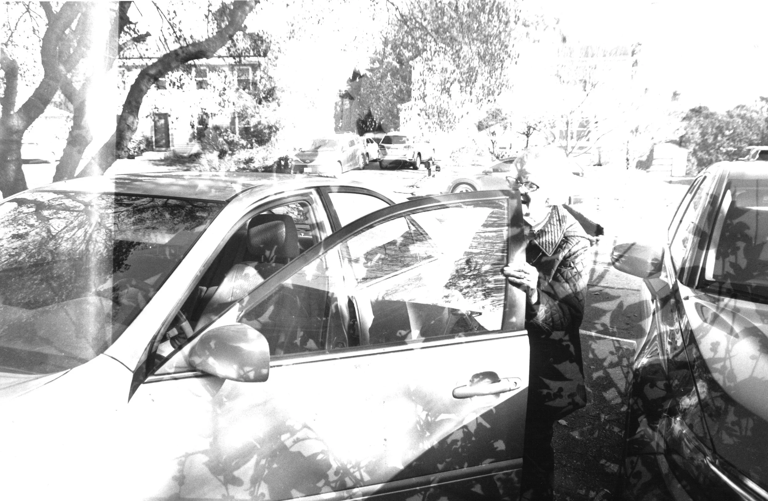 A person opening the front passenger door of a car parked in an outdoor parking lot, with trees and other cars in the background.