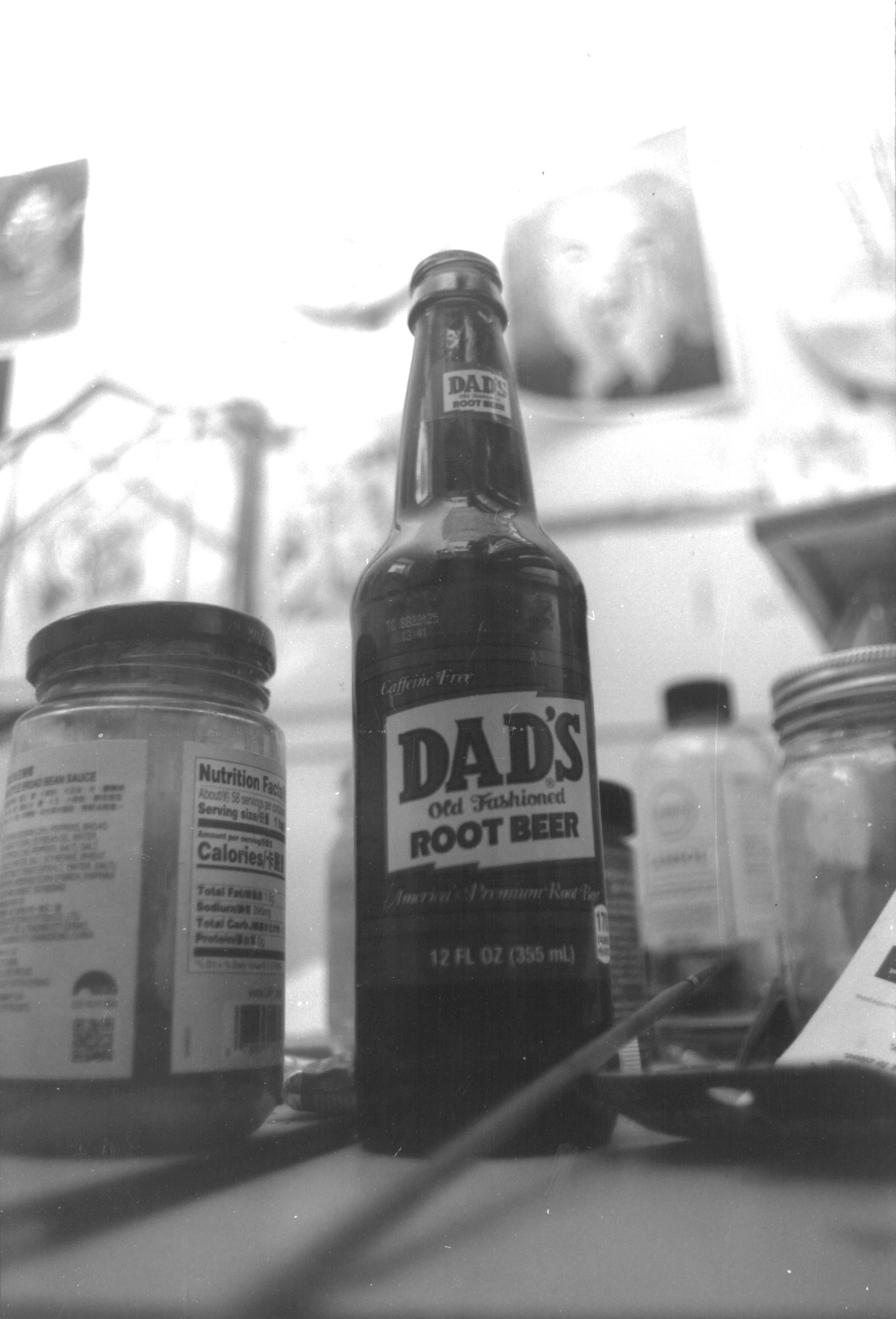 Black and white photo of a bottle labeled 'Dad's Old Fashioned Root Beer' on a cluttered surface, surrounded by jars and other objects.