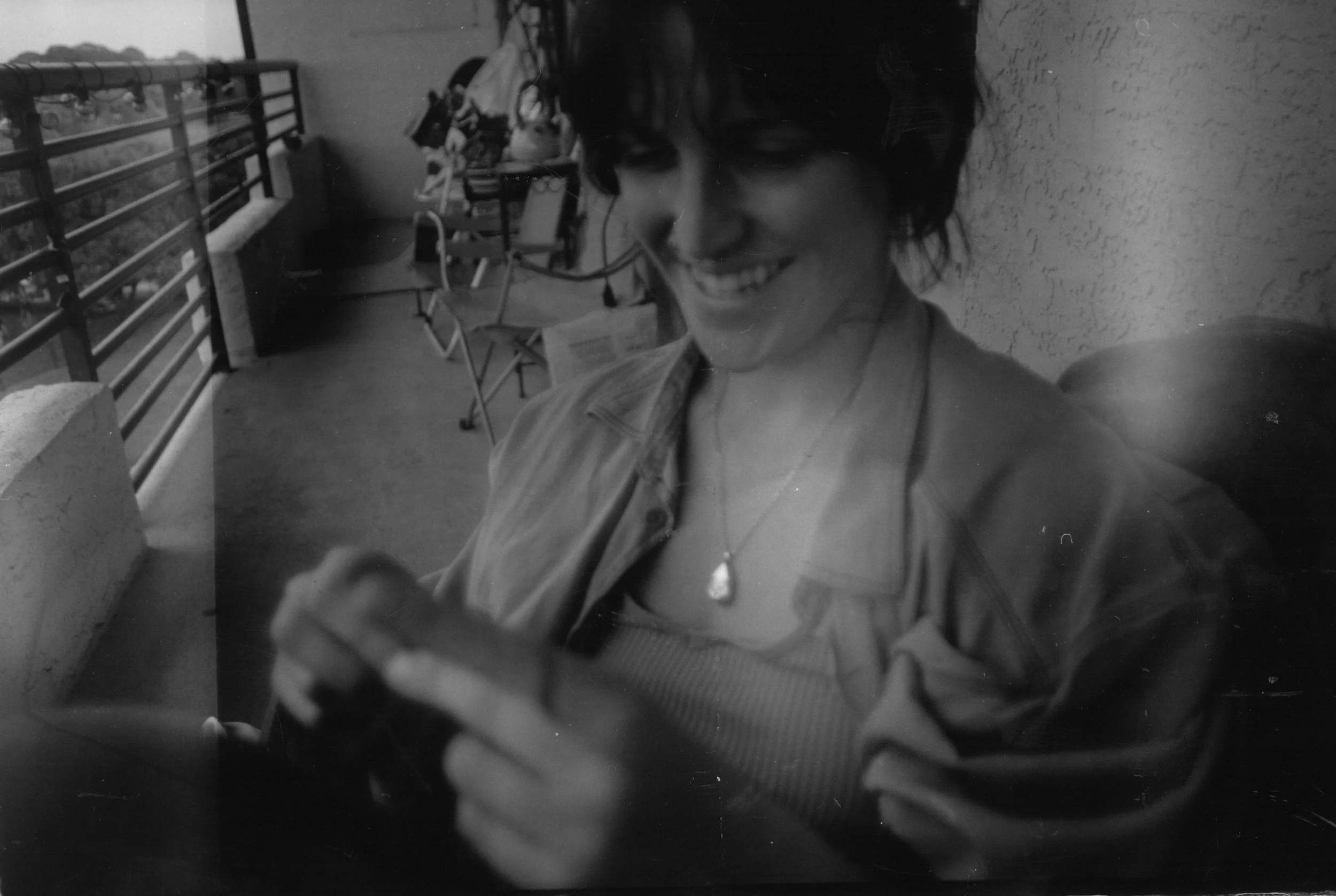 A woman with short dark hair, smiling and looking at her phone, sitting outdoors on a balcony with chairs and a railing in the background.