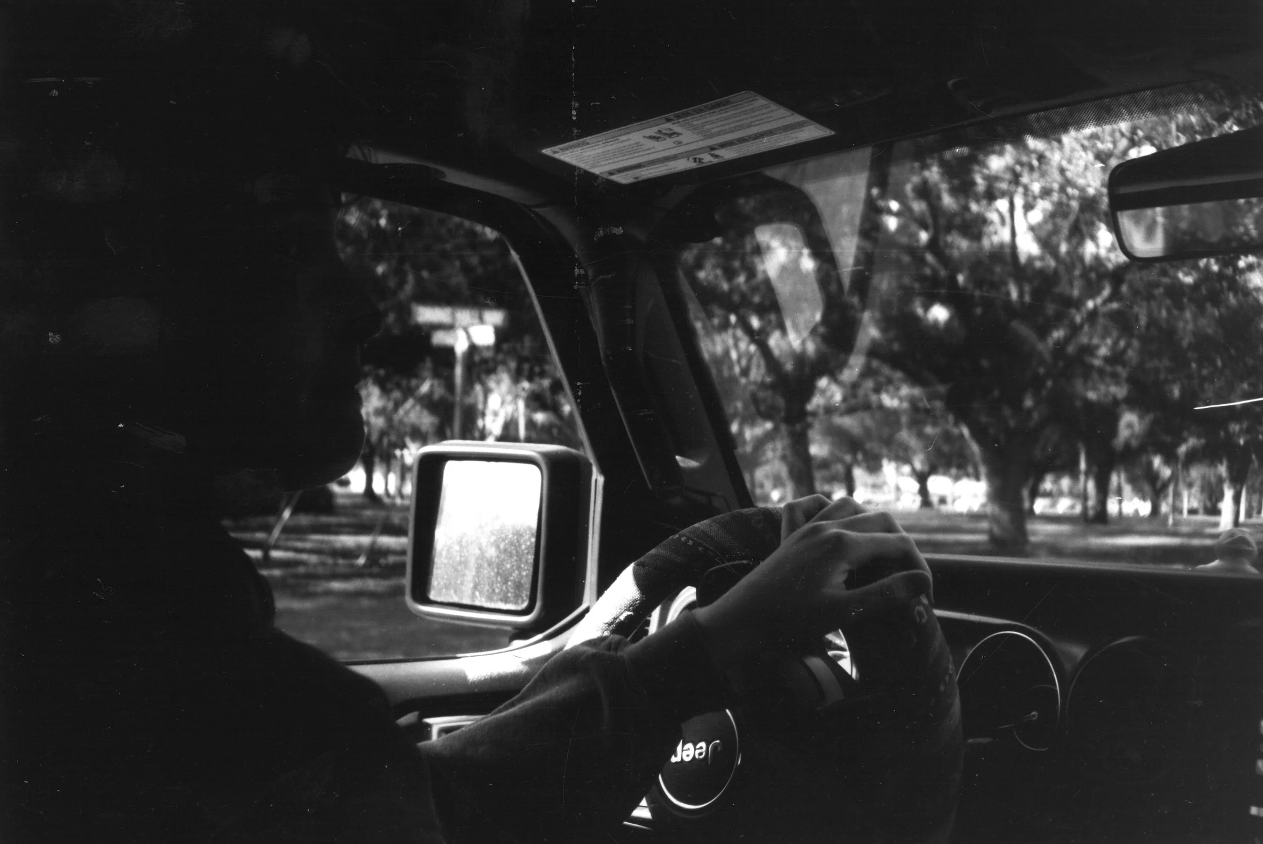 Black and white photograph of a person driving a Jeep vehicle, viewed from the passenger side. The driver's hand is on the wheel, and the view outside shows trees and a park-like setting.