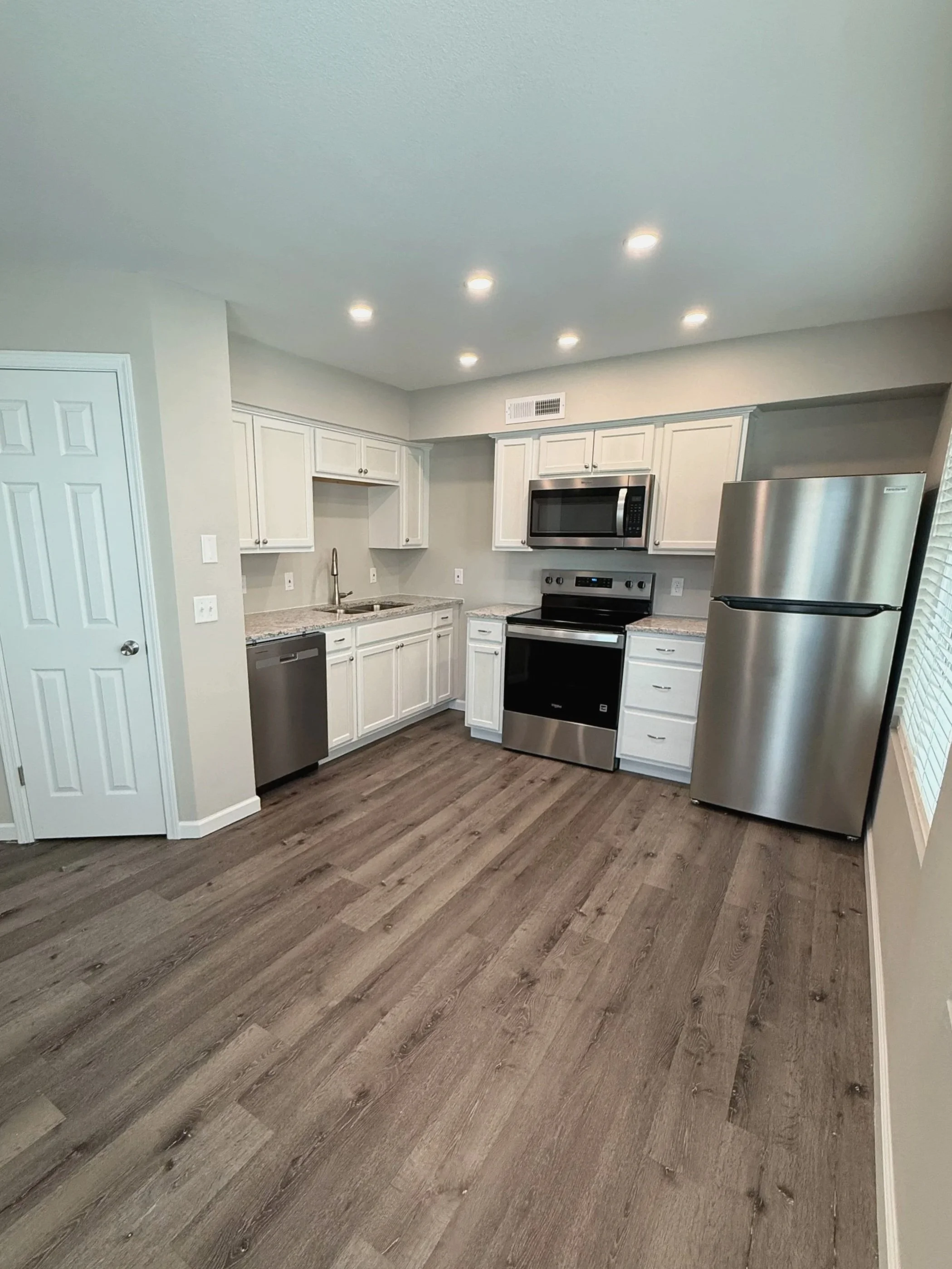 Modern kitchen with white cabinets, stainless steel appliances including a refrigerator, oven, microwave, and dishwasher, with wood-style vinyl plank flooring and recessed lighting.