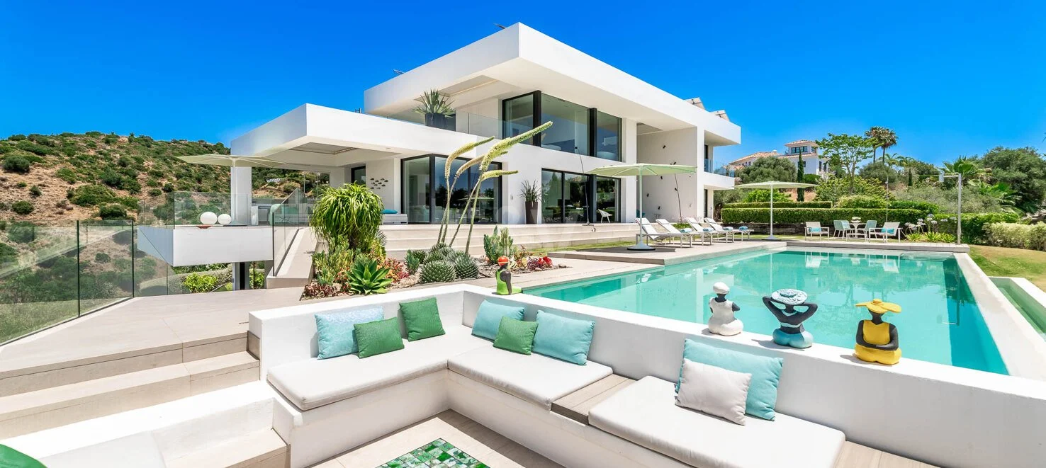 Modern white house with large glass windows and outdoor patio, swimming pool with lounge chairs, and desert landscape with hills in the background.