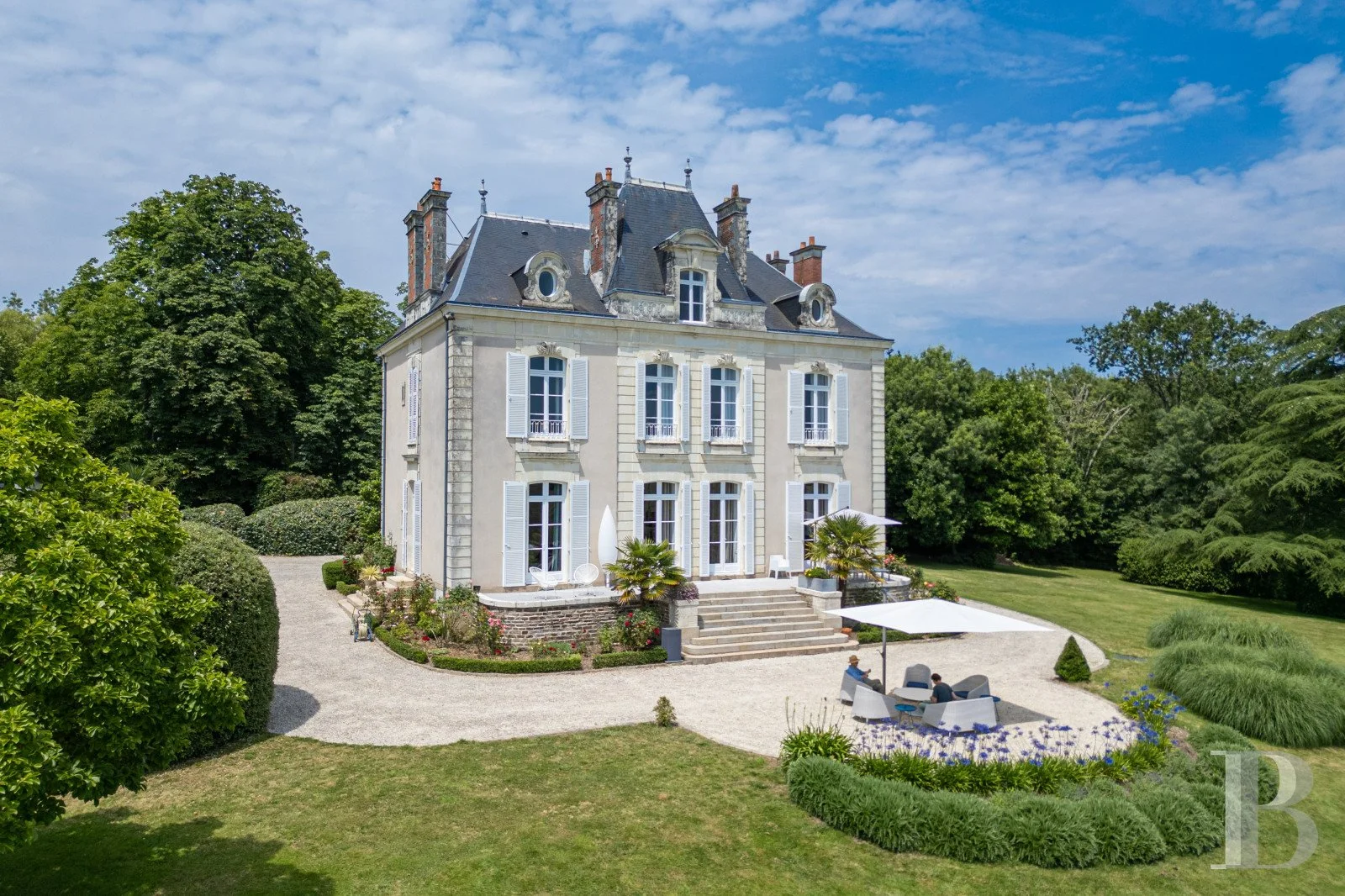 A large, elegant mansion with gray roofing and white shutters, surrounded by lush green trees and landscaped gardens, with a gravel patio featuring outdoor seating with umbrellas.