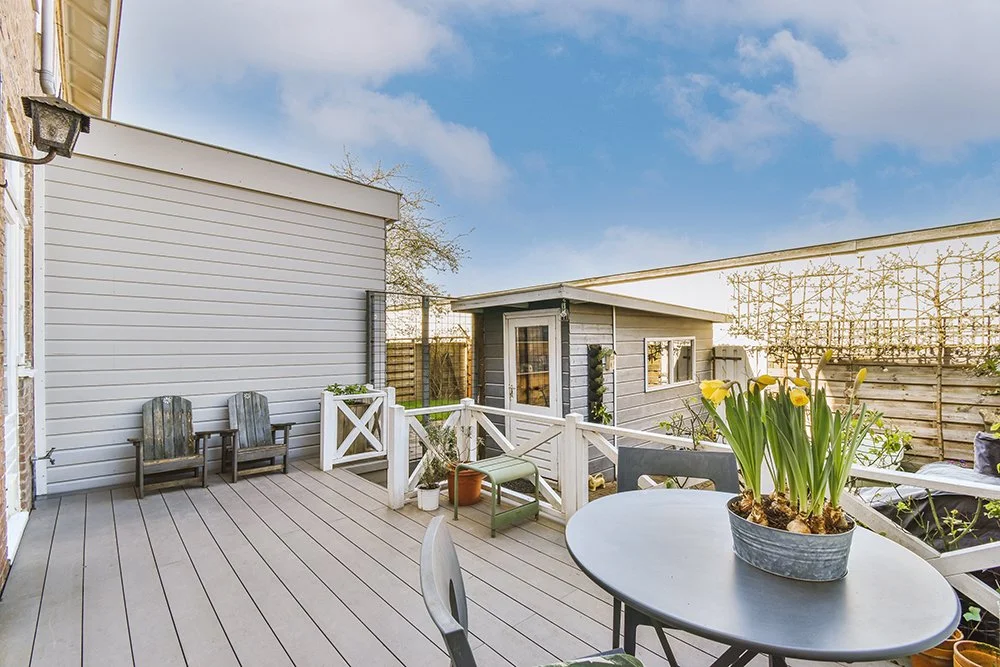 A backyard patio with a round table holding a pot of yellow flowers, two wooden chairs, and a small gray building with a door and windows, surrounded by fencing and blue sky