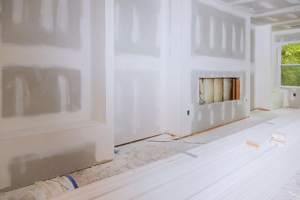 Interior of a house under construction with drywall walls, a window, and construction materials on the floor.