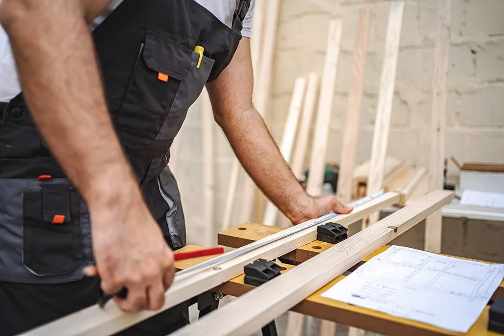 A person working on construction, measuring a piece of wood with a ruler in a workshop with framing and construction plans on the table.