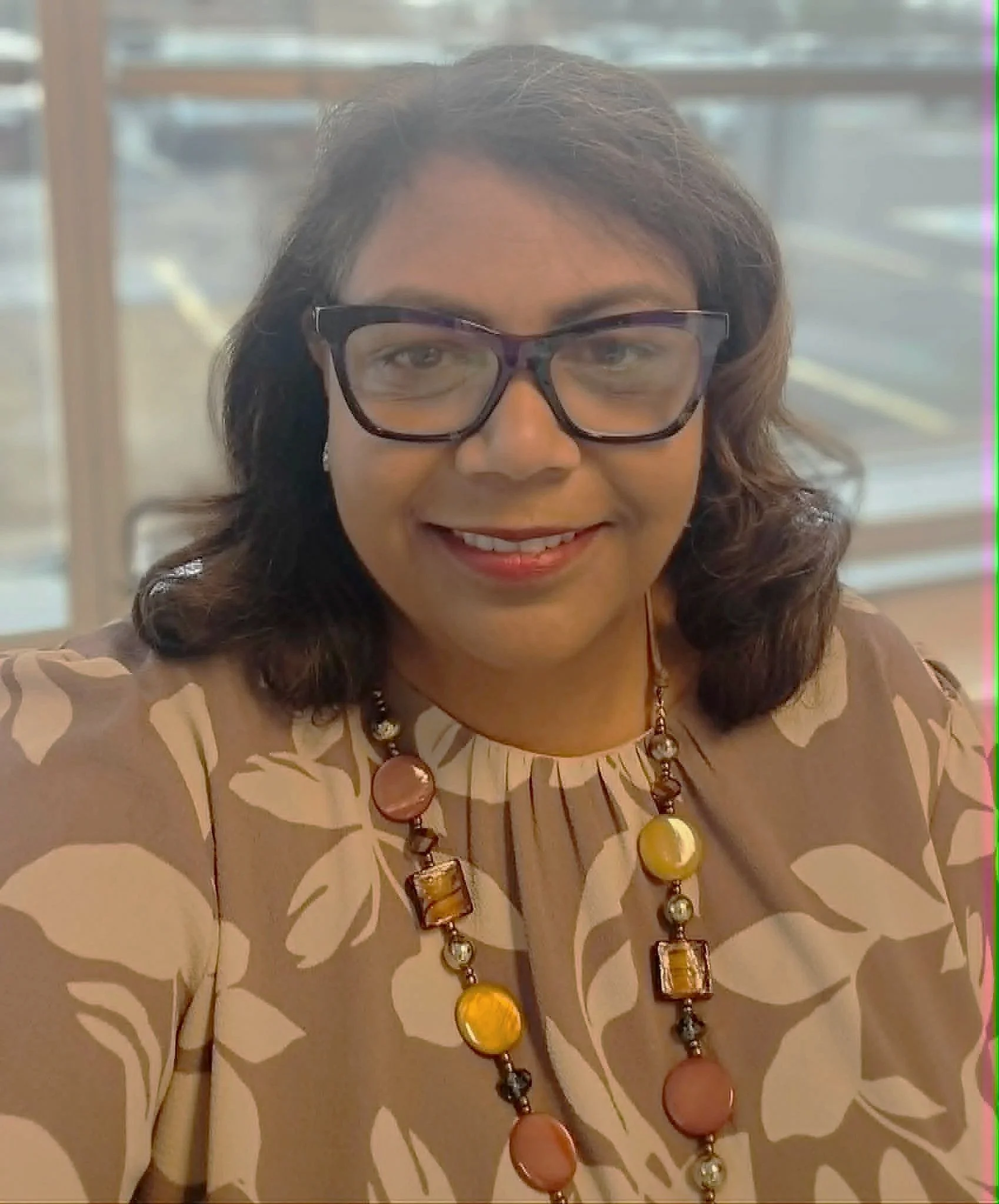 A close-up of a woman with glasses and earrings smiling, wearing a beige patterned top with brown, orange, and yellow accents, with a blurred background of a window and indoor setting.