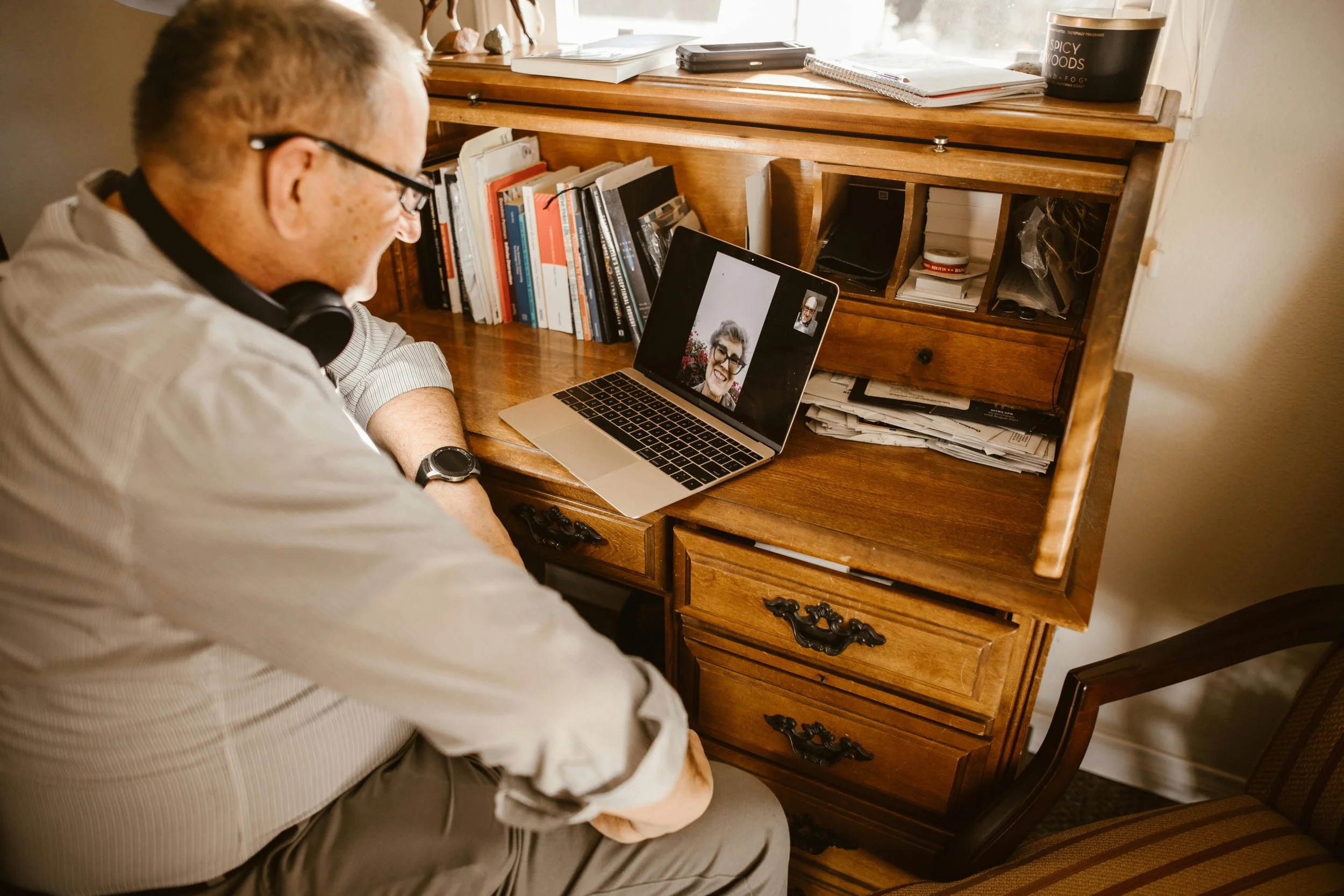 Man sitting at a wooden desk engaging in a video call on a laptop, with a bookshelf and cluttered workspace behind him.