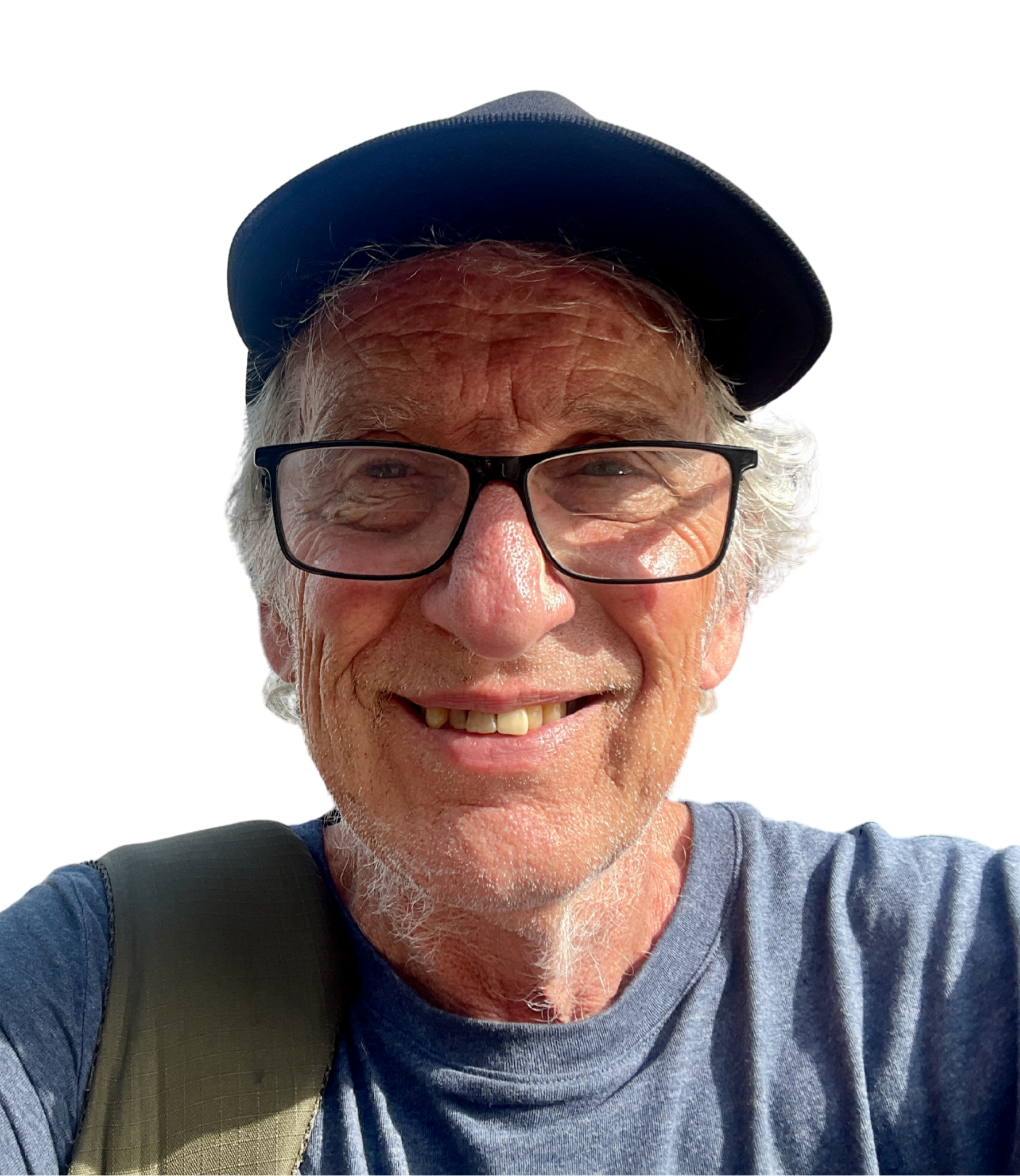 Close-up selfie of a smiling older man wearing glasses, a cap, and a backpack, with a bright background.