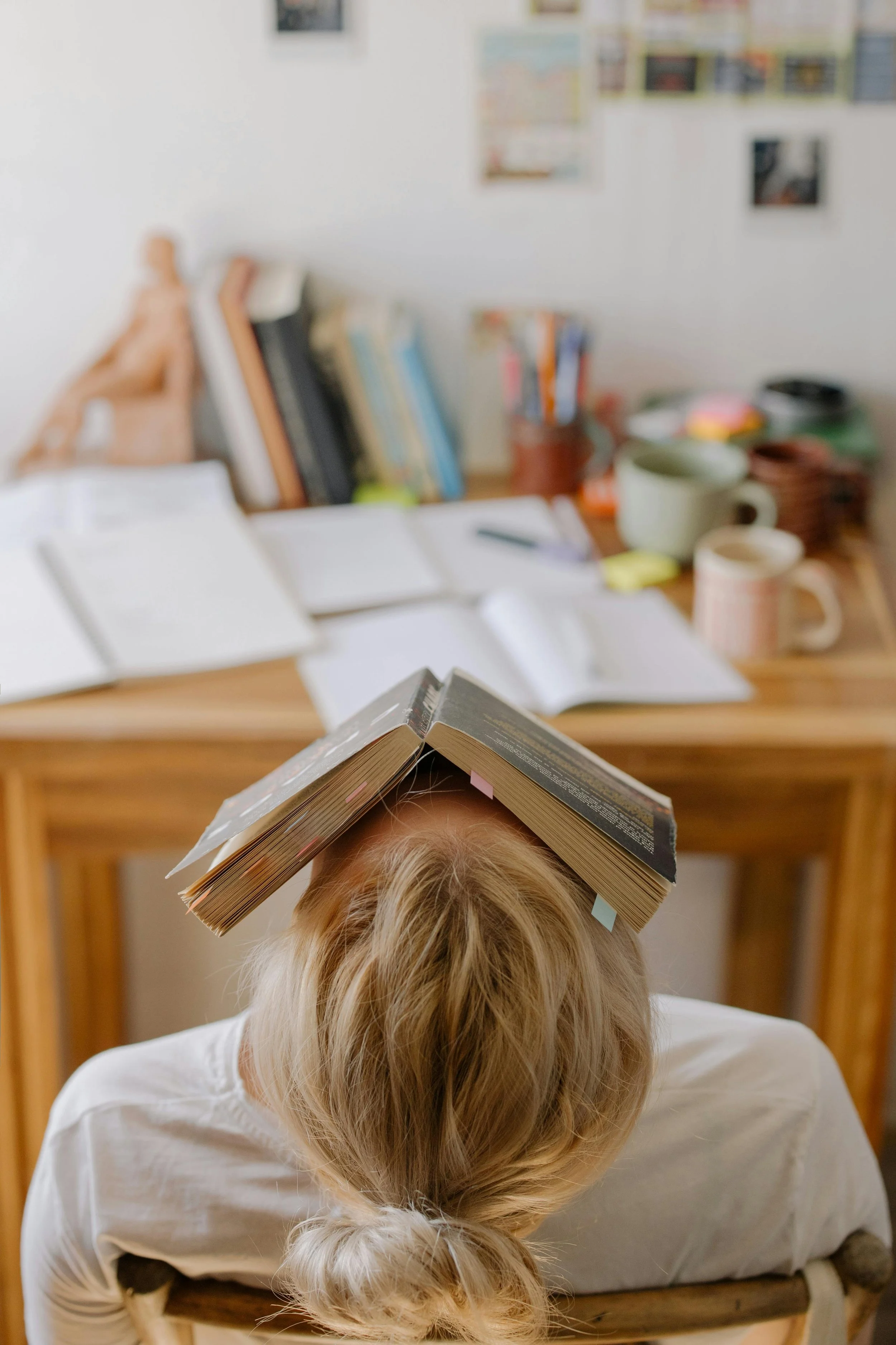 A person with blond hair tied back, sitting at a desk with a book balanced on their head. The desk is cluttered with open books, notebooks, mugs, and various office supplies.