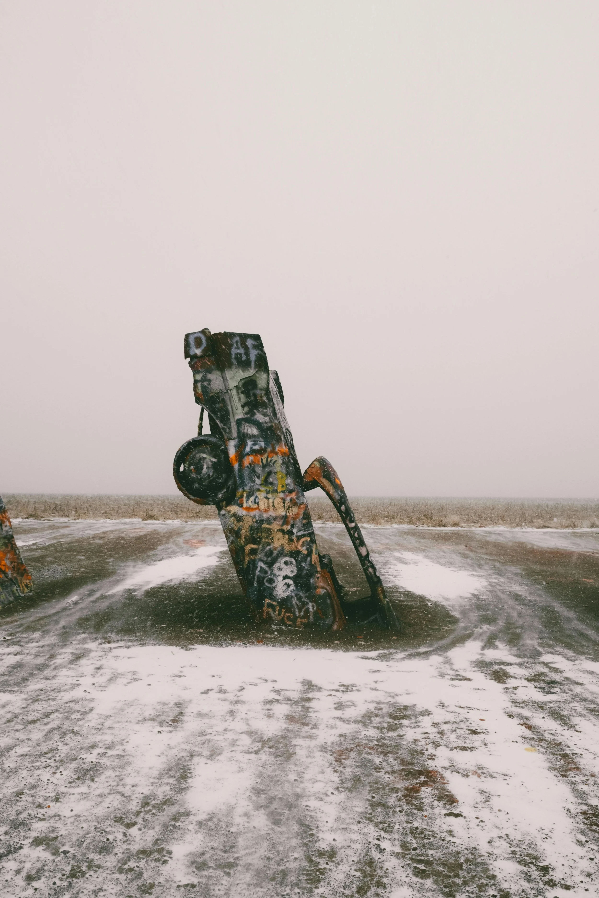A large sculpture resembling an old-fashioned vending machine partially submerged in the sand and water, covered in colorful graffiti, on a foggy beach.