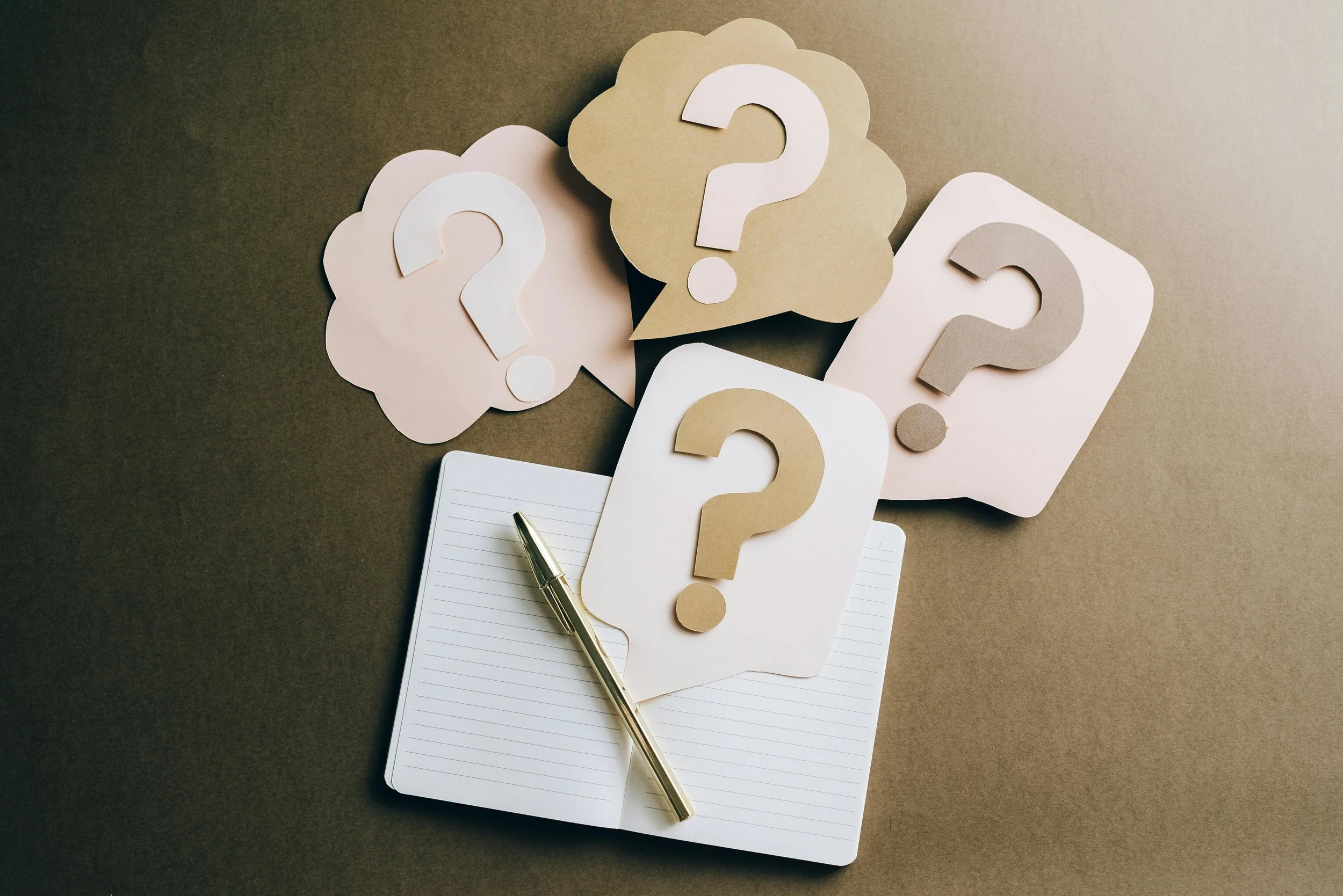 A notebook with a pen, surrounded by paper cutouts of speech bubbles with question marks in various colors, on a brown background.
