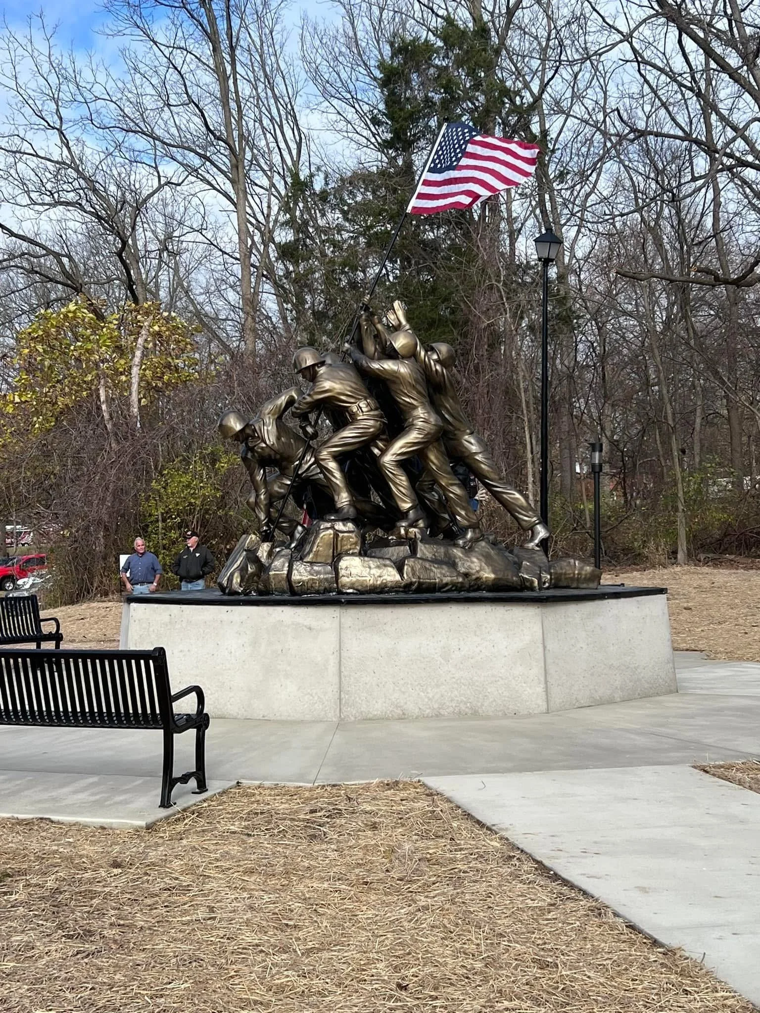 Bronze statue of soldiers raising the American flag, located on a stone base in a park with leafless trees and benches nearby.