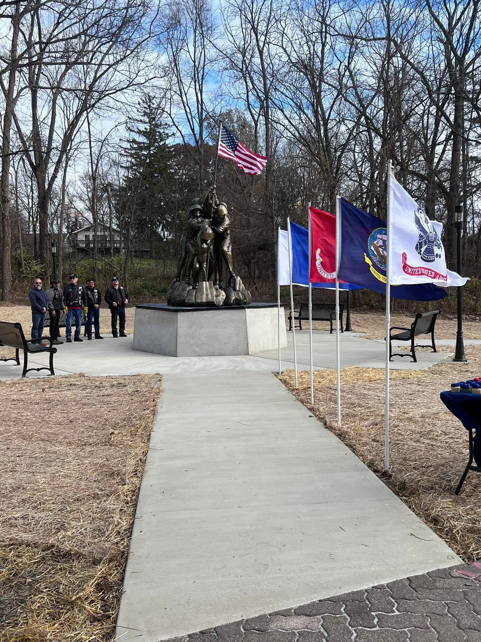 A memorial park featuring a statue of soldiers raising a flag, surrounded by flags, benches, and a group of people paying respects amid leafless trees.