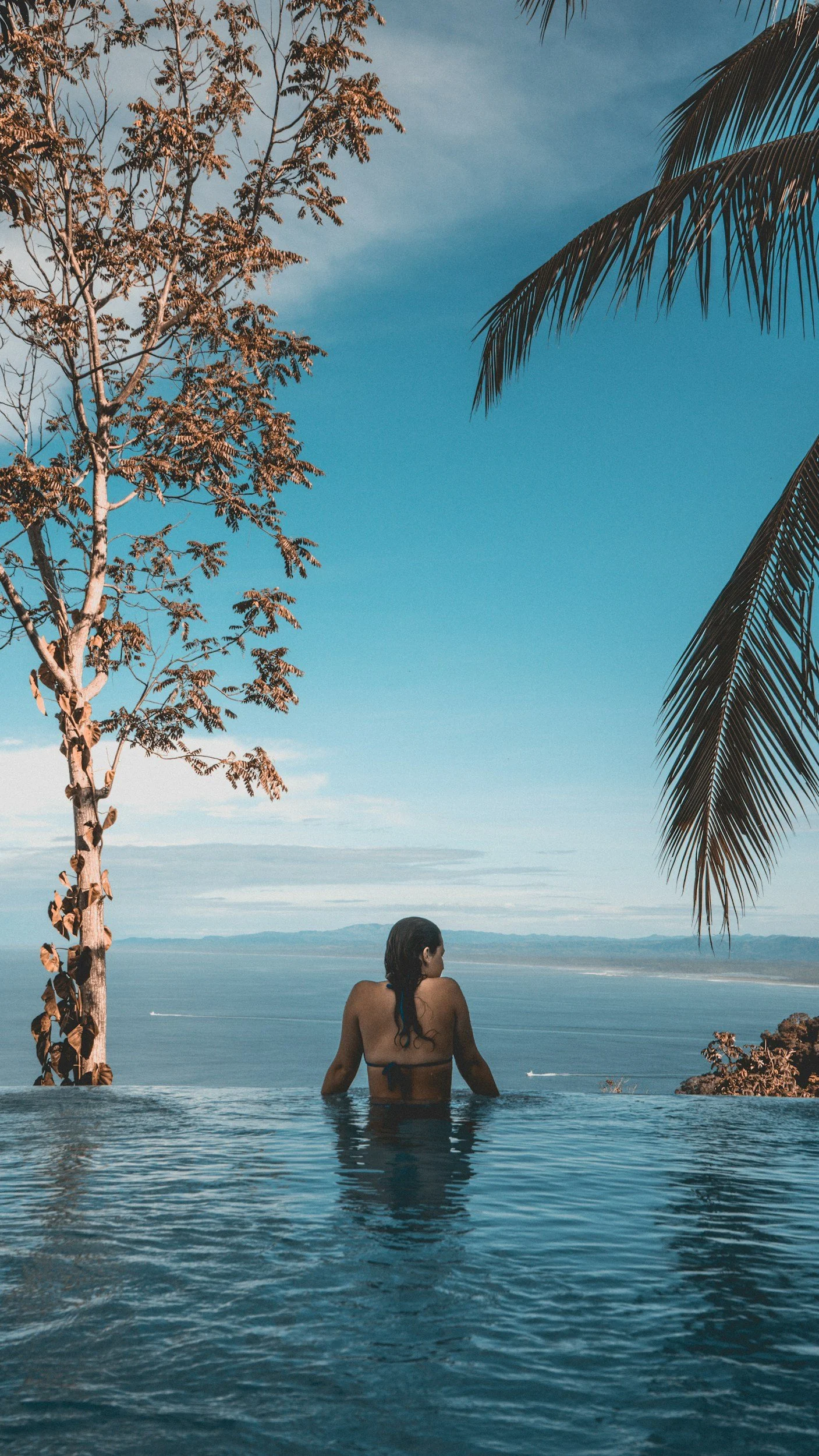 A woman in a bikini sitting in a swimming pool, overlooking a body of water with mountains in the distance, surrounded by palm trees and a partly cloudy sky.