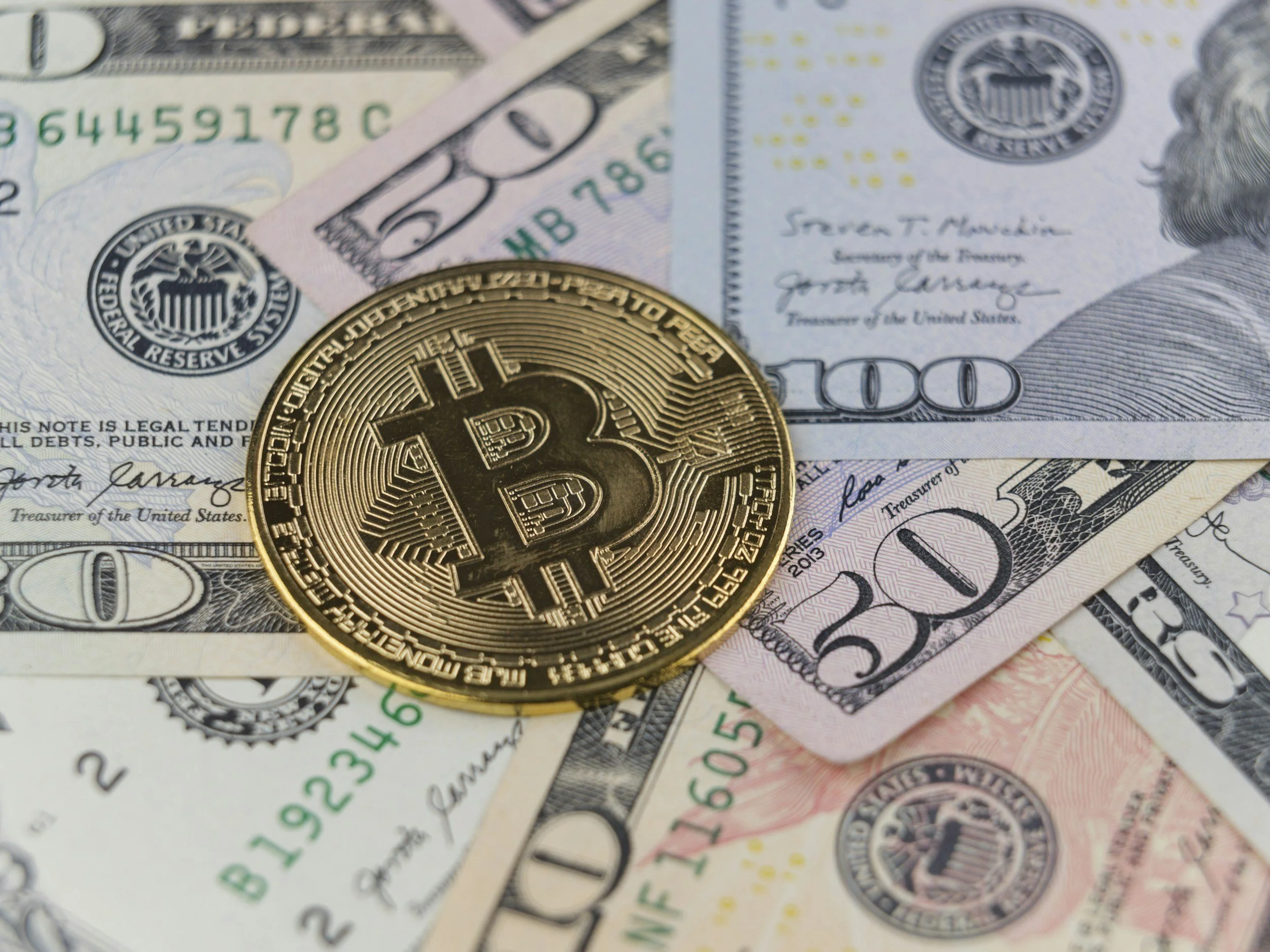 A physical Bitcoin coin placed on US dollar bills, including $100, $50, and $20 bills.