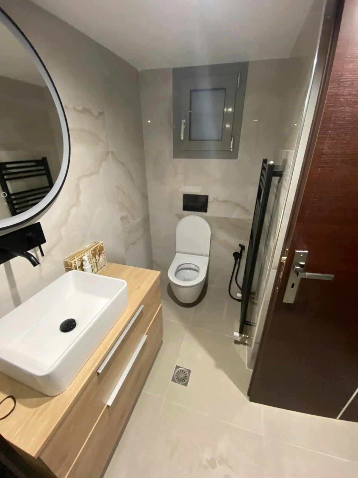 Ανακαίνιση μπάνιου με πλακάκι τύπου μάρμαρμο, μαύρα εντοιχισμένα μπιντέ, πετσετοκρεμάστρα, καζανάκι και μπαταρία. Ο νιπτήρας είναι επικαθήμενος σε ξύλινο έπιπλο.