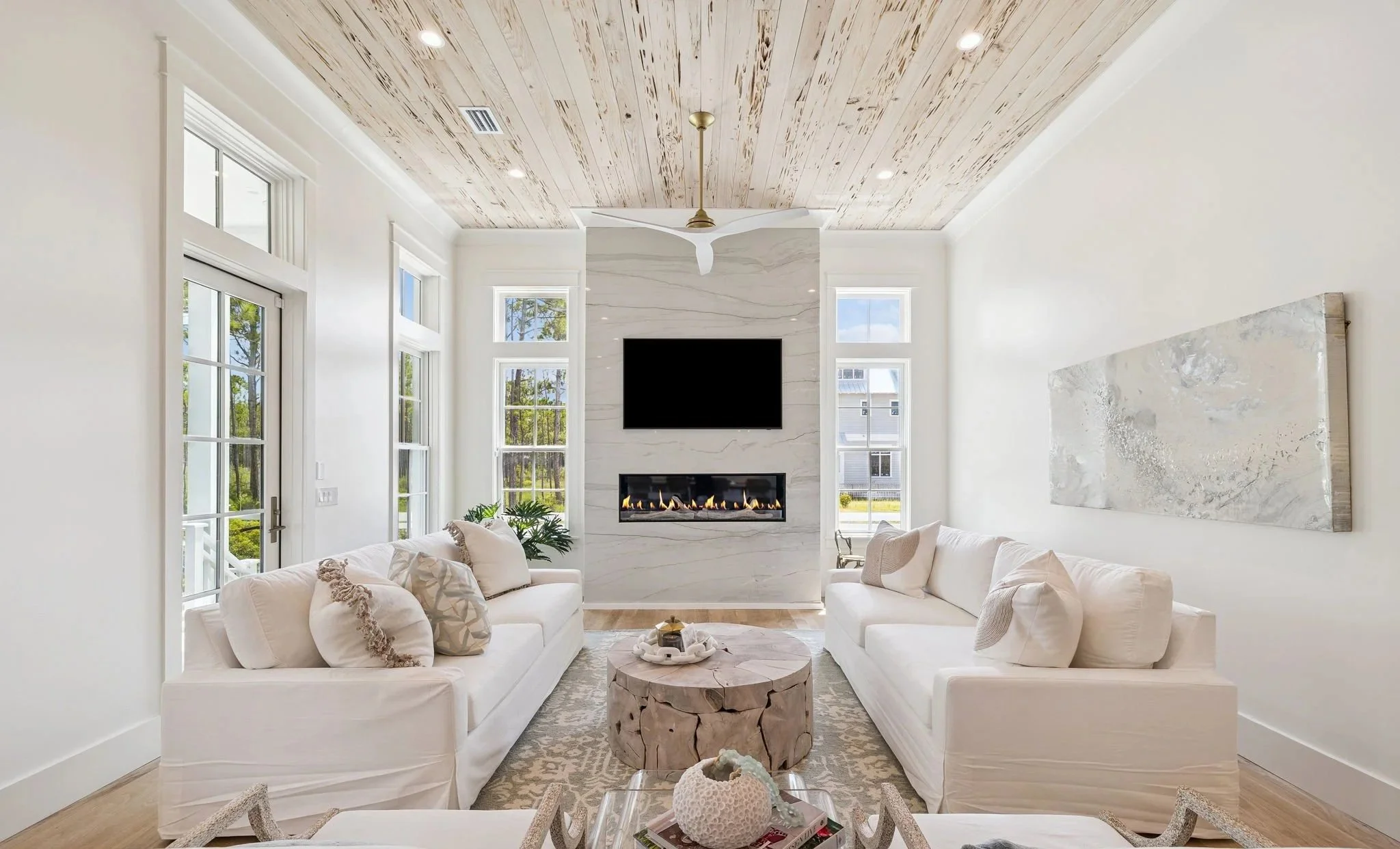 Bright living room with two white sofas, a wooden coffee table, large windows, a marble fireplace, a TV mounted above the fireplace, and a wooden ceiling with a ceiling fan in 30A.