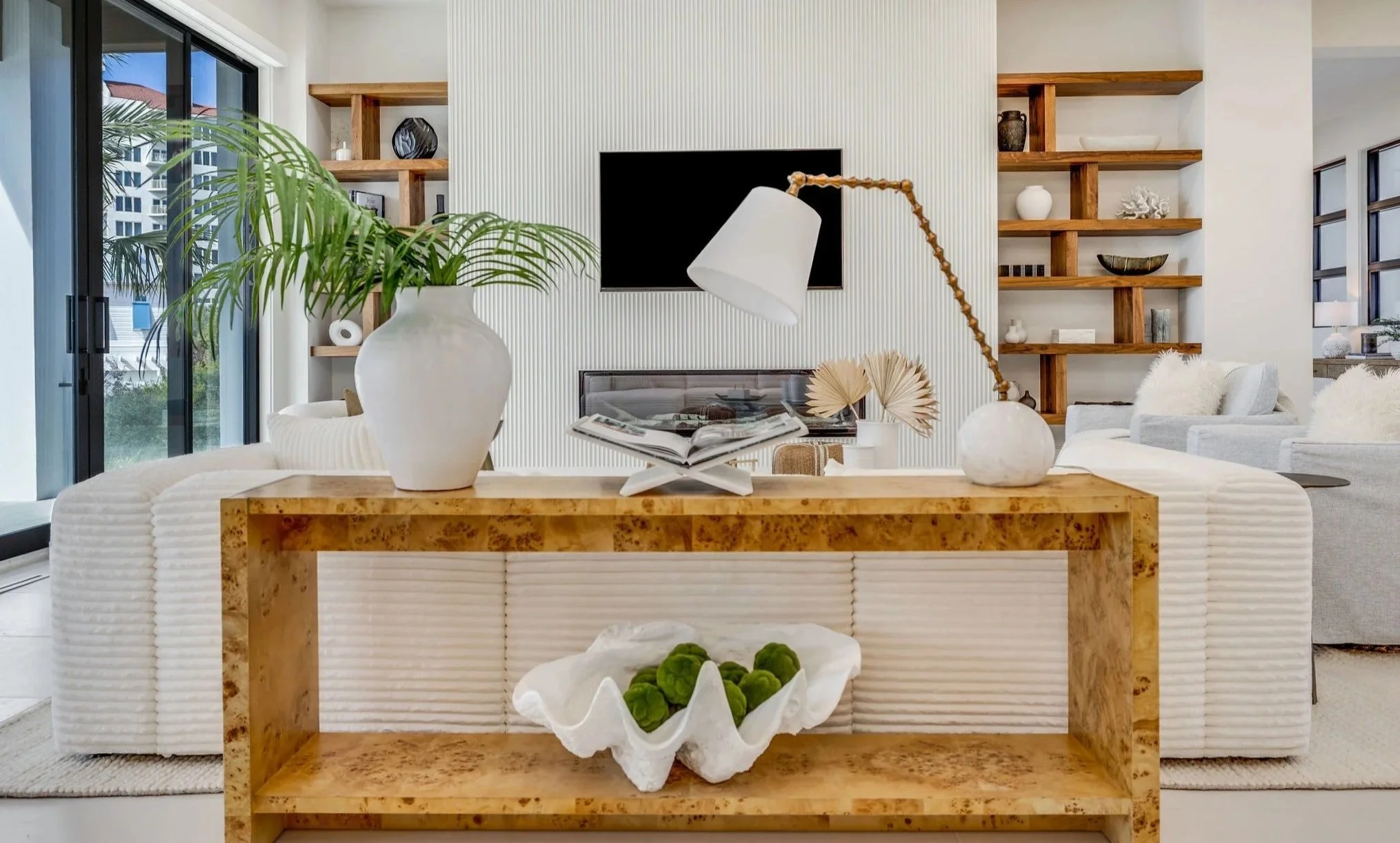 Modern living room with a wooden console table holding a large white vase with a green plant, a white lamp, and an open book. Behind the console, a sofa with white cushions. In the background, a textured white wall with wall-mounted shelves and a tel