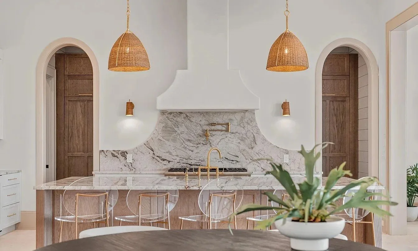 Modern kitchen with white walls, marble backsplash, wooden cabinets, gold fixtures, and two wicker pendant lights. A white potted plant is on the foreground table.