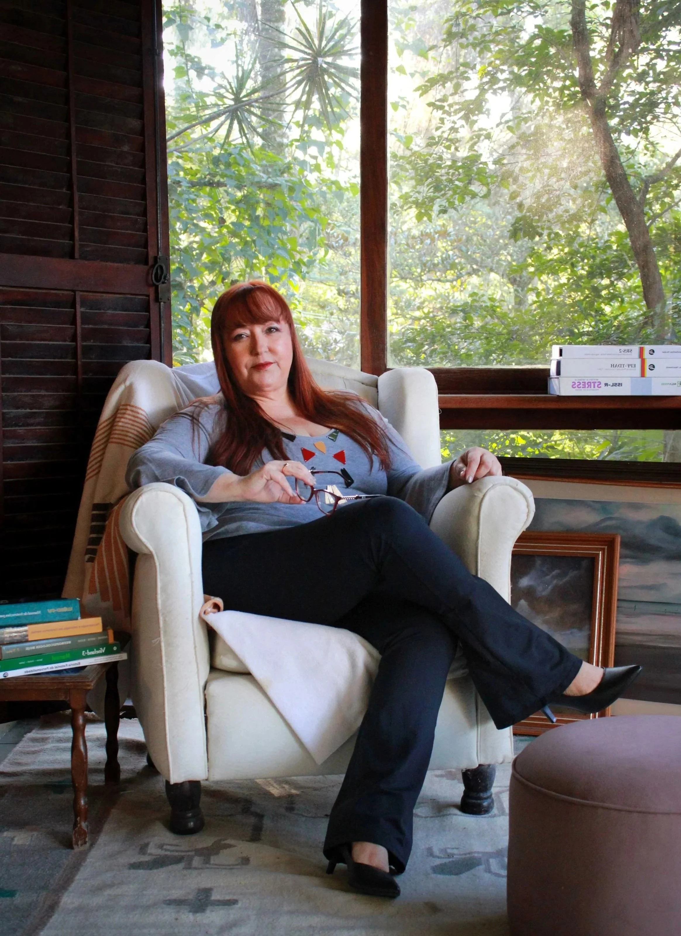 Mulher com cabelo ruivo, usando camiseta cinza, sentada em um sofá de veludo bege, com uma coluna de livros ao lado esquerdo, na frente de uma janela grande com vista para árvores verdes e céu claro.
