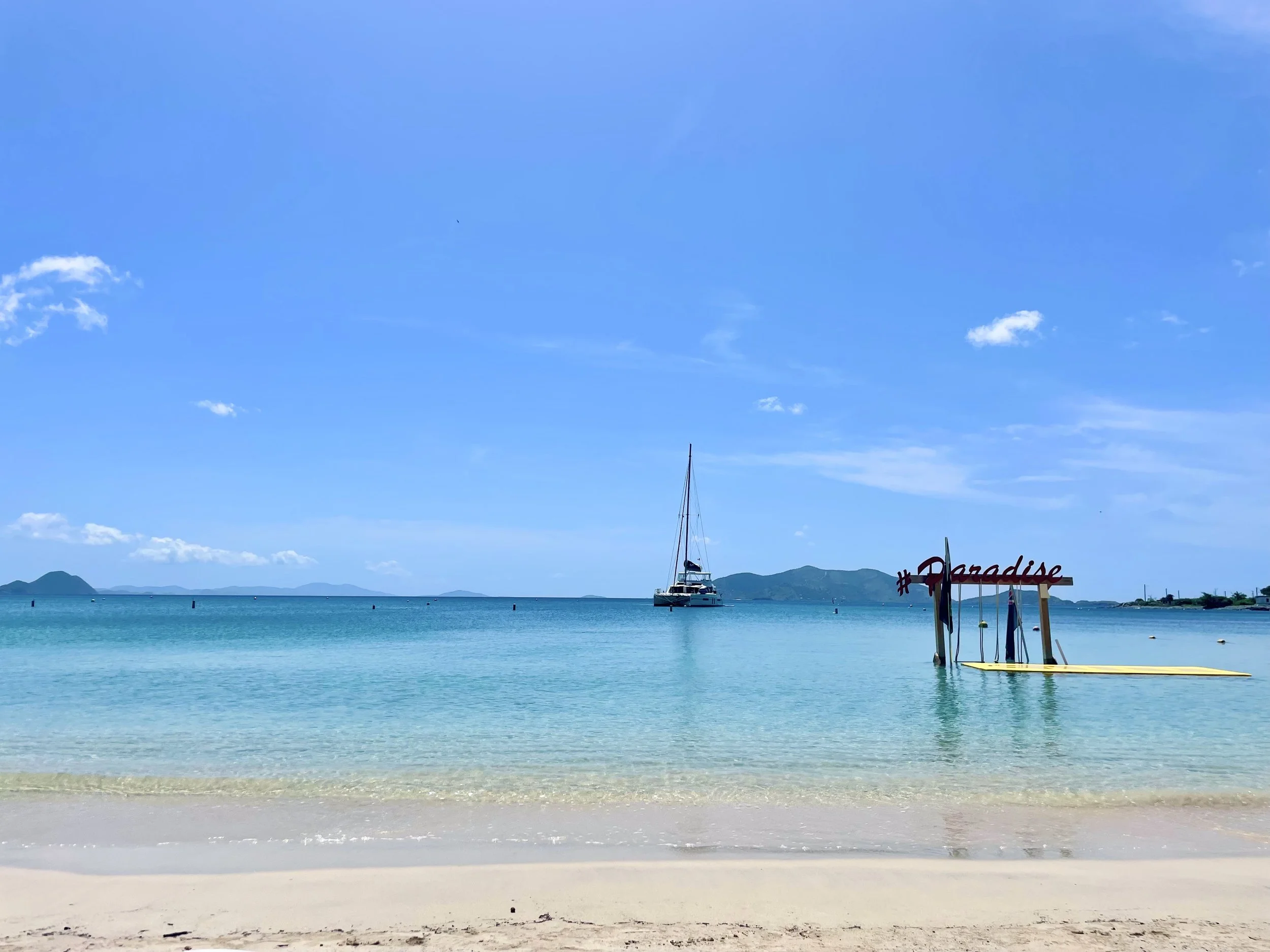 A peaceful beach scene with clear turquoise water, a sailboat in the distance, and a yellow paddleboard with a red sign reading '#Paradise' near the shore, against a backdrop of distant mountains and a blue sky with some clouds.