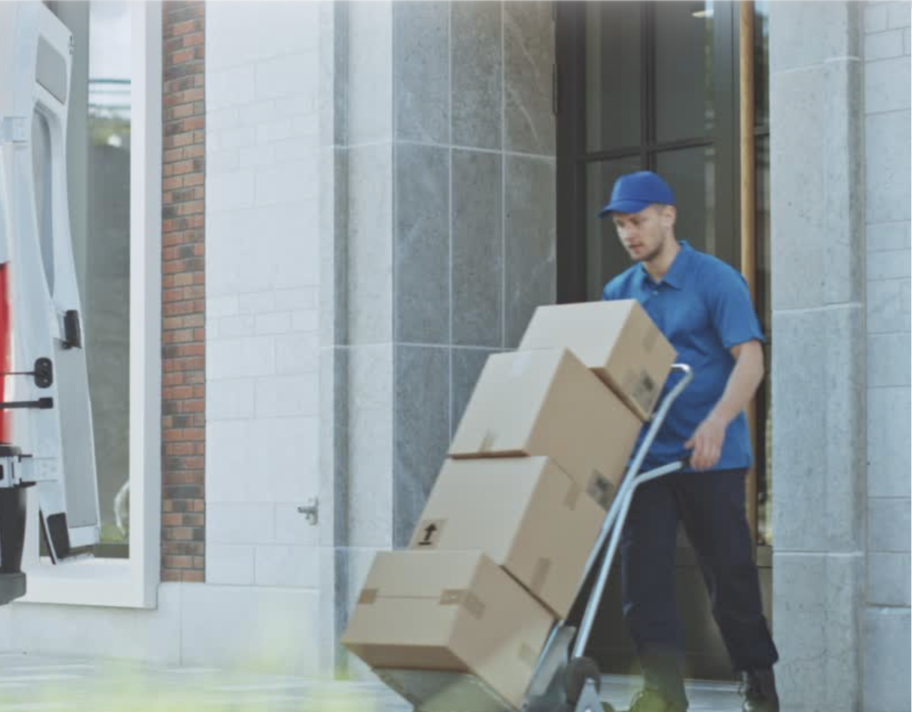 A delivery person in a blue uniform and cap is unloading boxes from a hand truck outside a building.