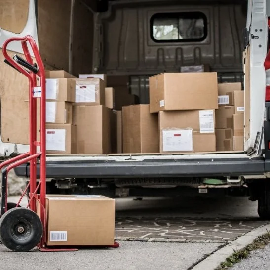 Open delivery van filled with cardboard boxes on the floor and on shelves, with a red hand truck in front of the van